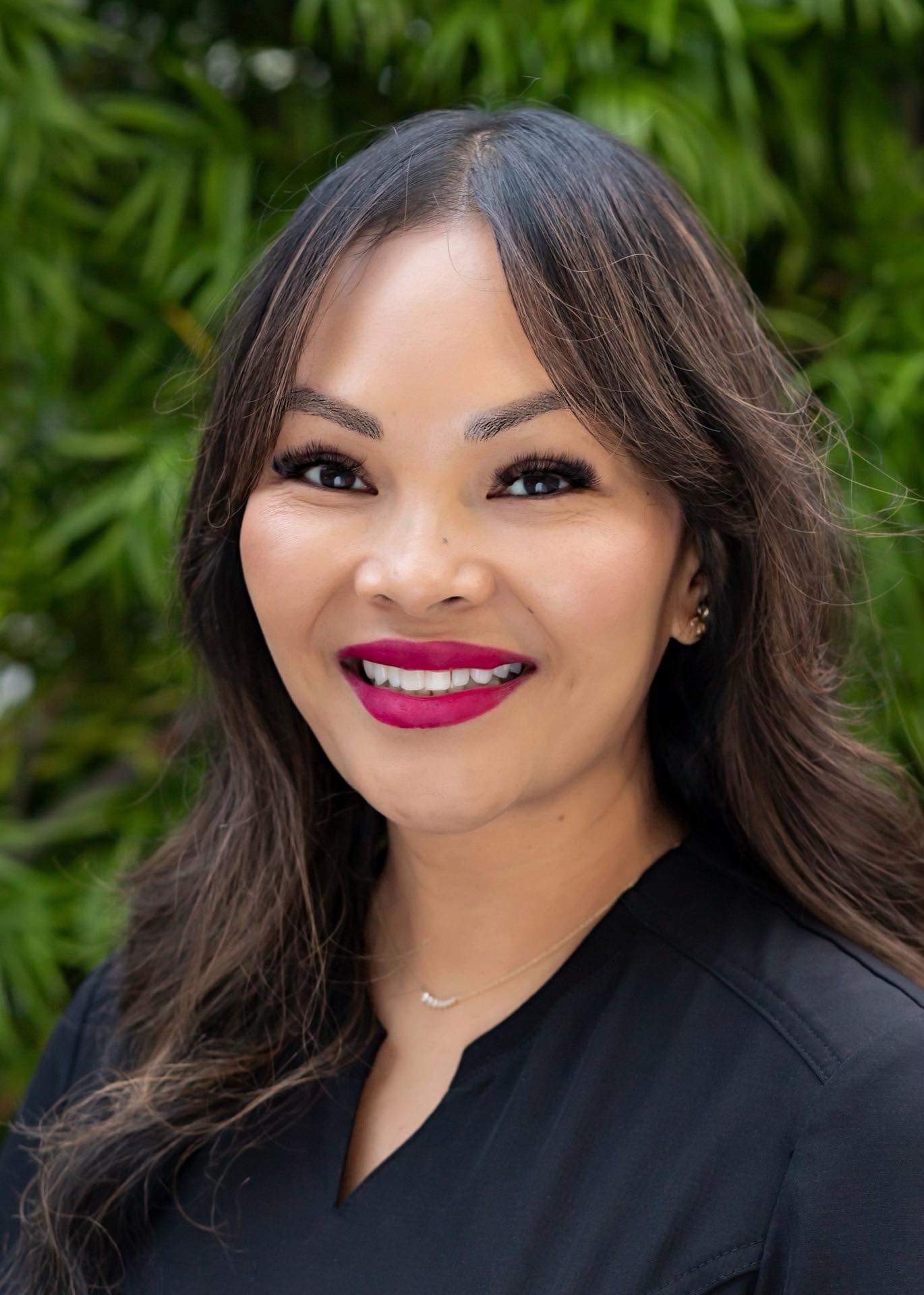 A woman with long brown hair, wearing a black top, smiling with red lipstick, wearing earrings and a delicate necklace, standing outdoors in front of green foliage.