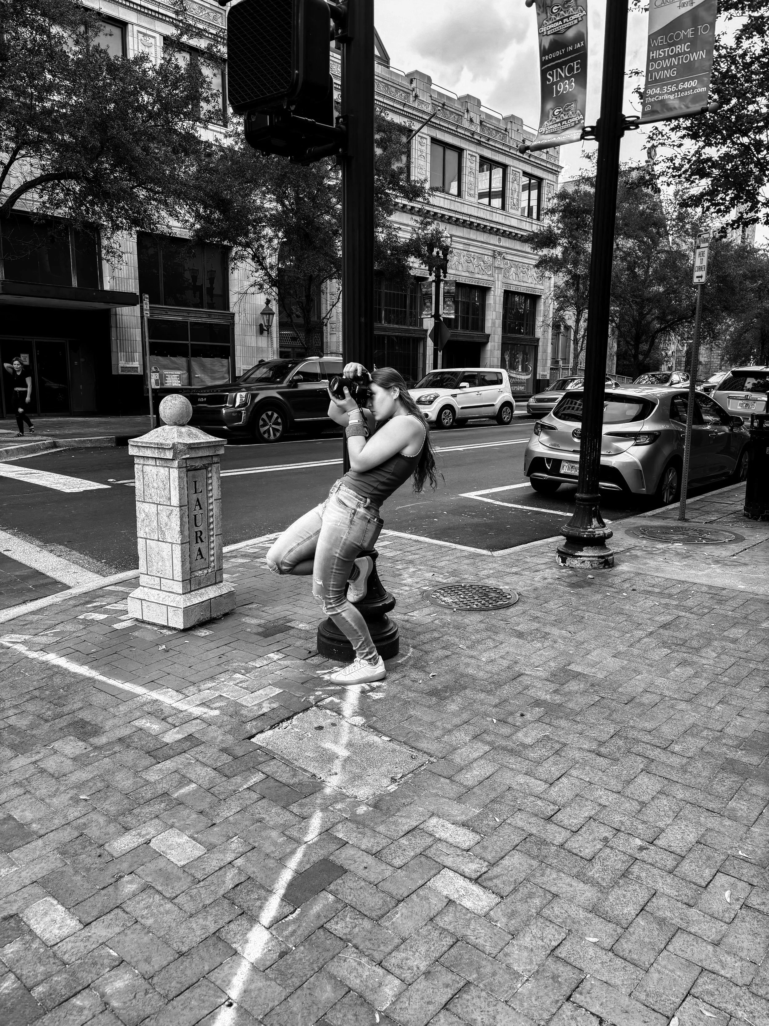 A woman taking a photograph on a city street corner with brick sidewalk, parked cars, and buildings in the background, in black and white.