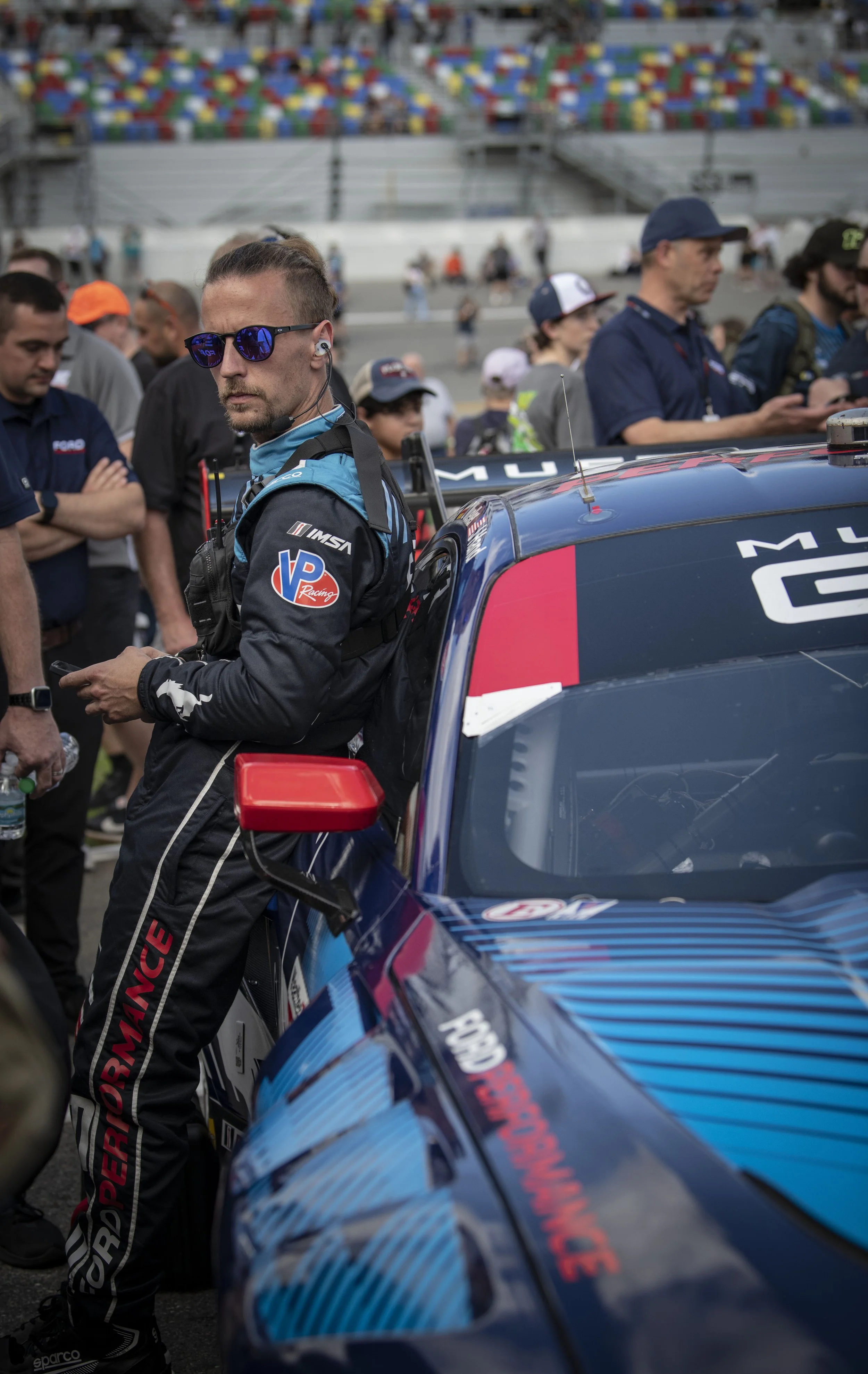 A race car driver in racing suit leaning against a blue and pink race car in the prep area of a racetrack, with multiple people and spectators in the background.