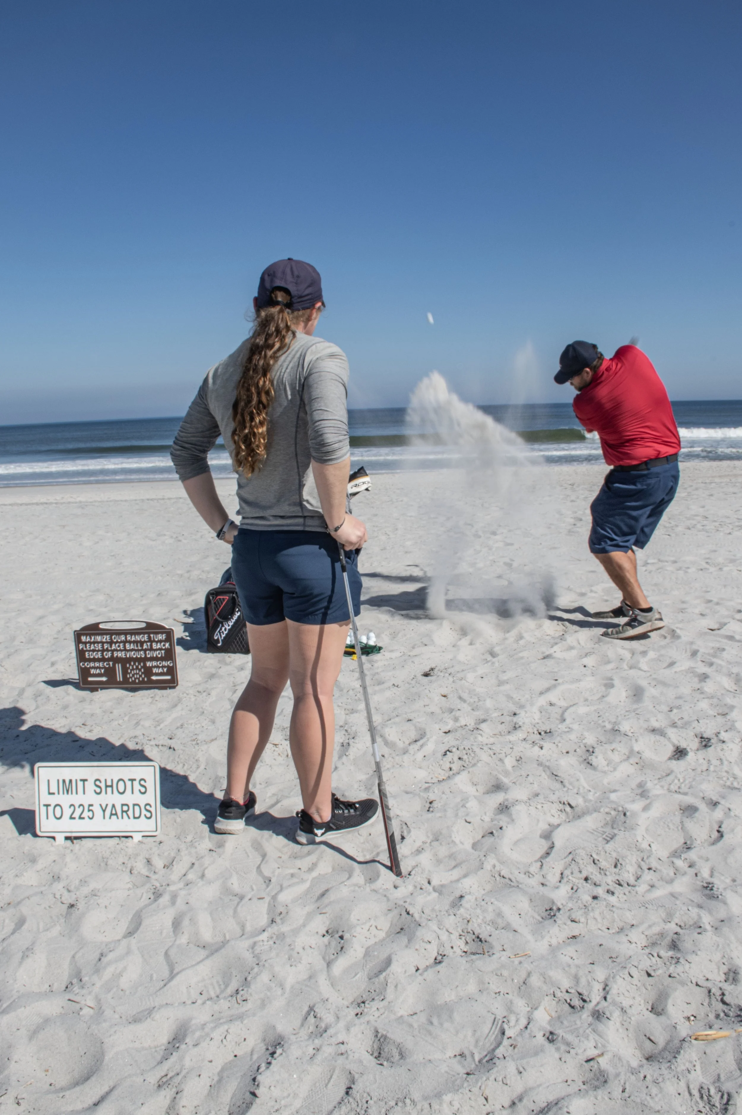 A man and a woman on the beach during the day, with the man preparing to hit a golf ball while the woman observes, holding a golf club; a sign states limits on shots and range rules, and the beach and ocean are visible in the background.