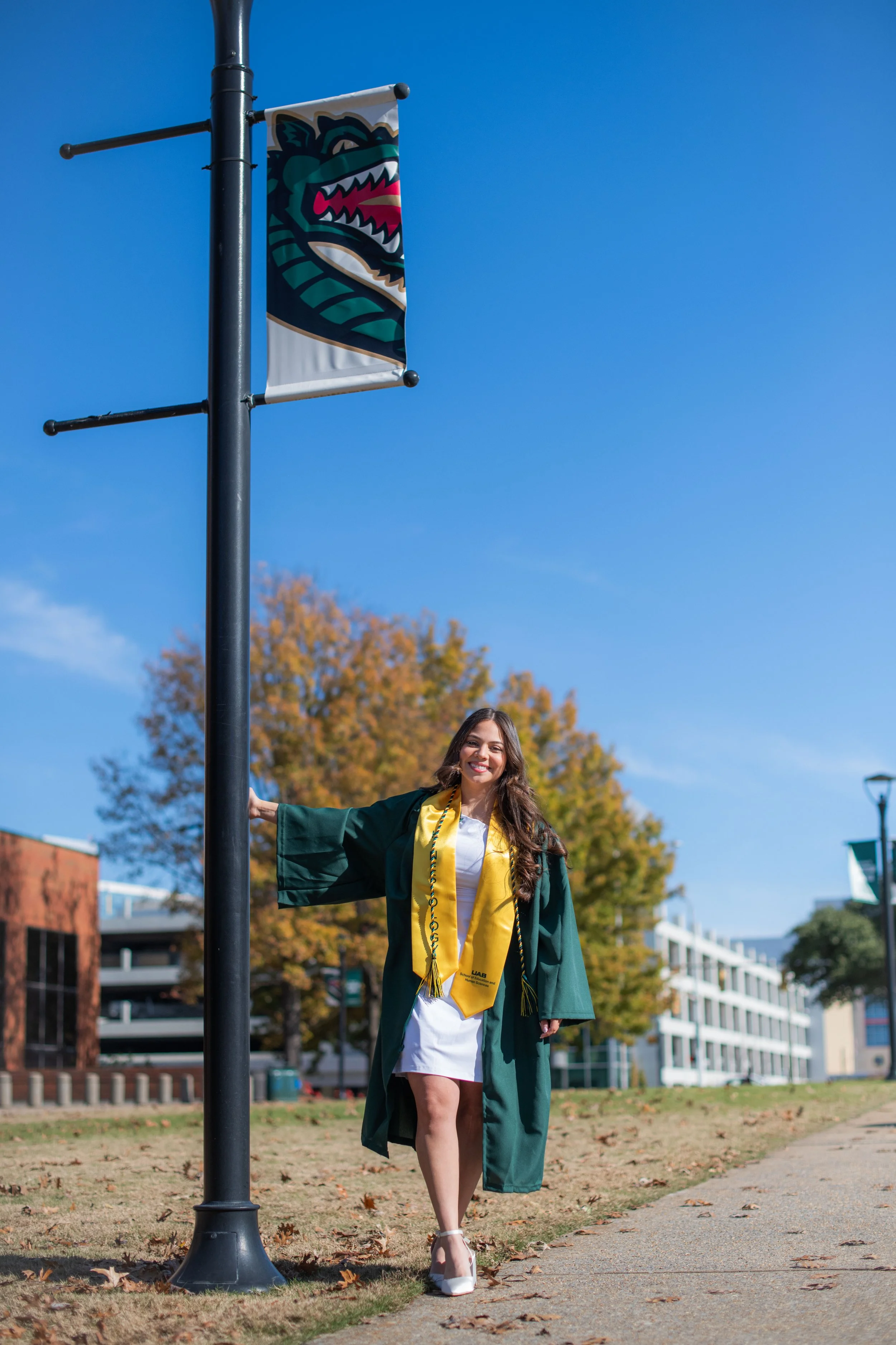 A young woman in a green graduation gown and yellow stole, smiling and posing outdoors on a sunny day, with autumn trees and buildings in the background.