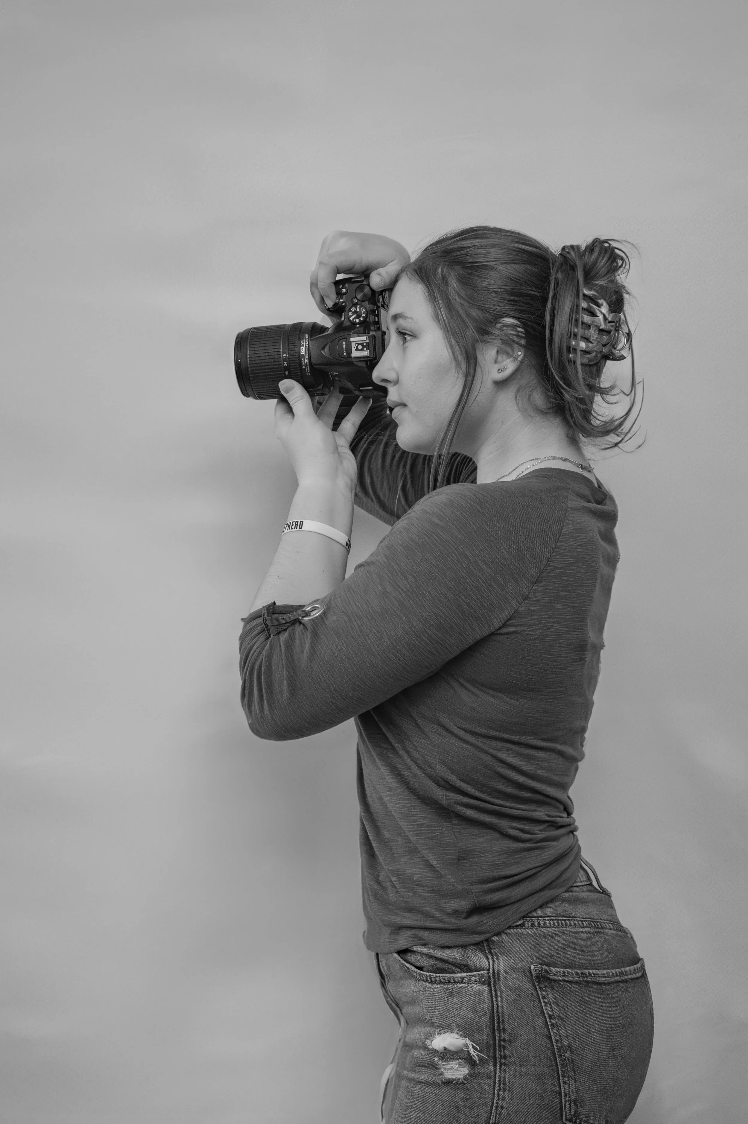 A woman with a ponytail hairstyle taking a photograph with a camera, standing sideways against a plain background.