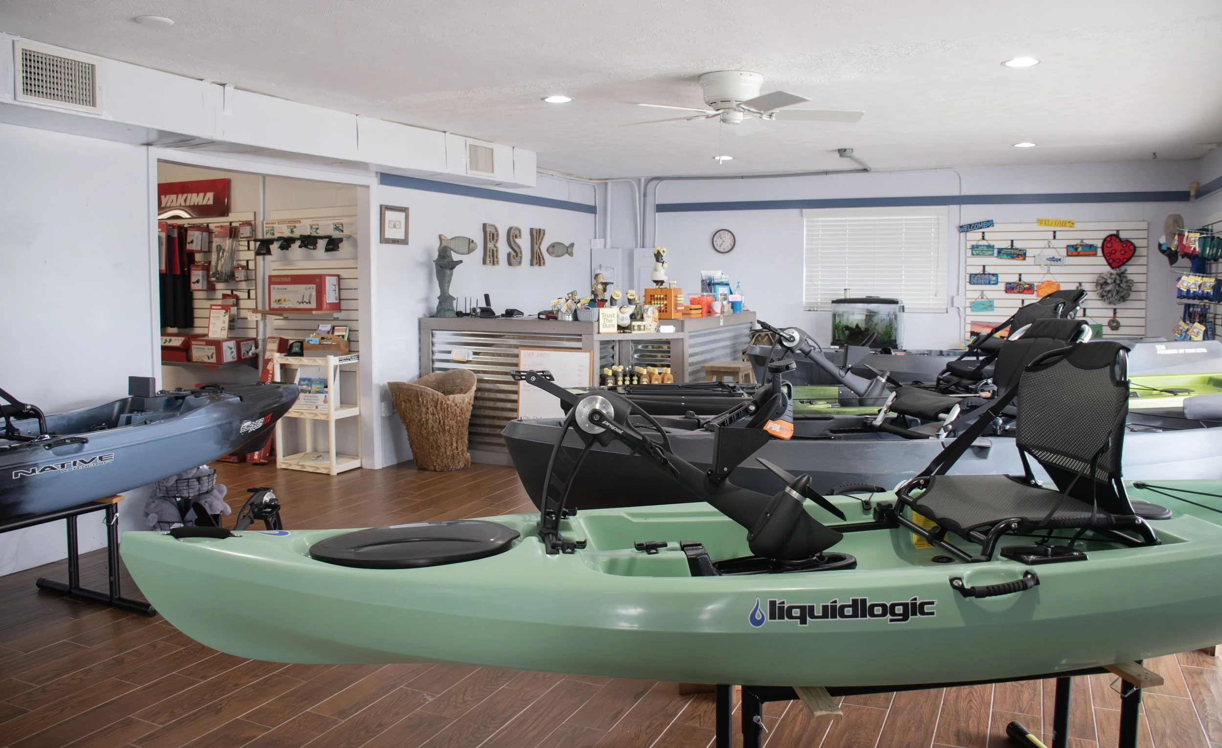 Indoor kayak retail store with multiple kayaks on display, including a green Liquidlogic kayak in the foreground, and various equipment and accessories on shelves and displays in the background.