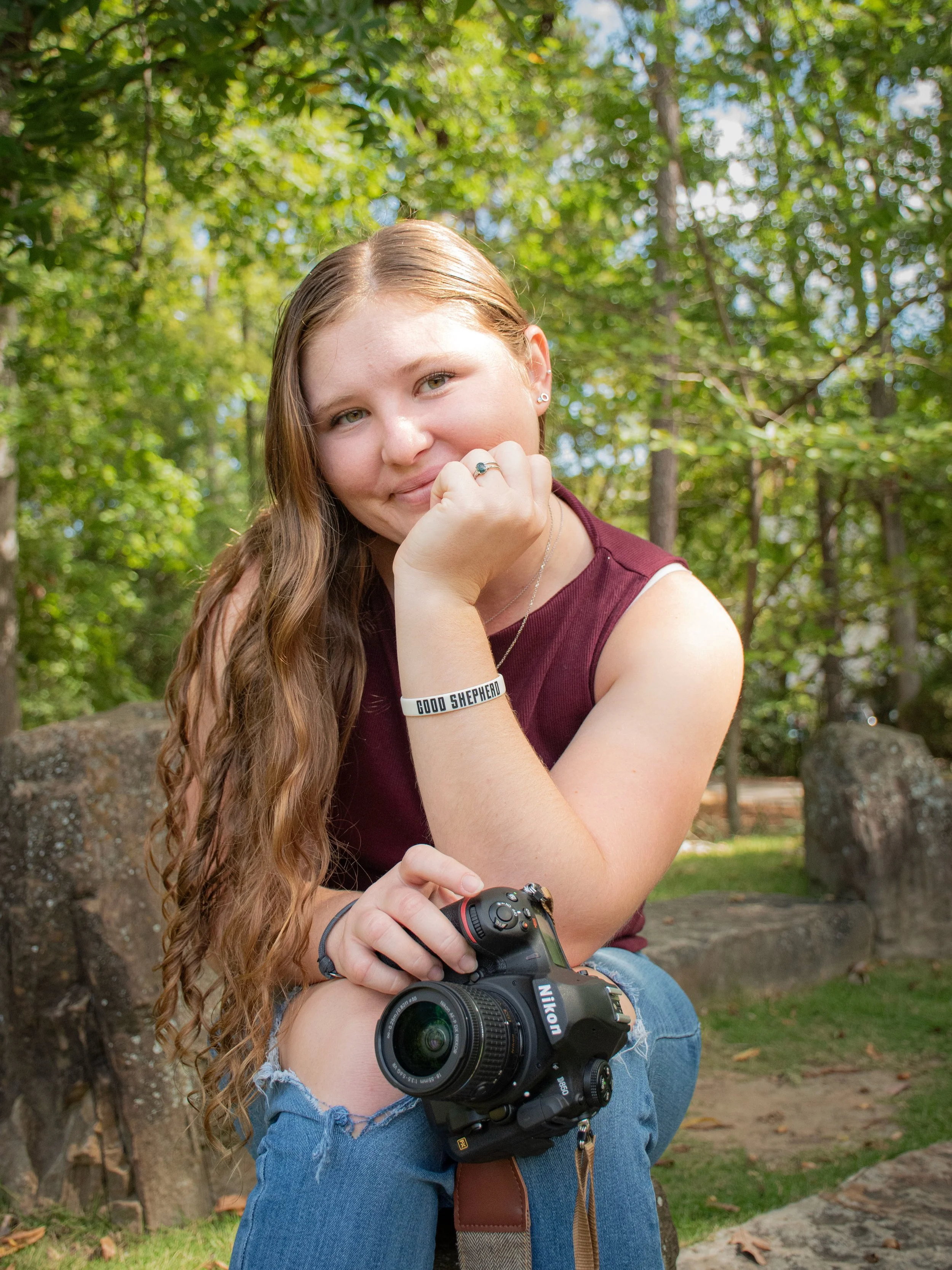 A young woman with long curly hair sitting outdoors on a stone bench in a wooded area, holding a camera on her lap, smiling at the camera.