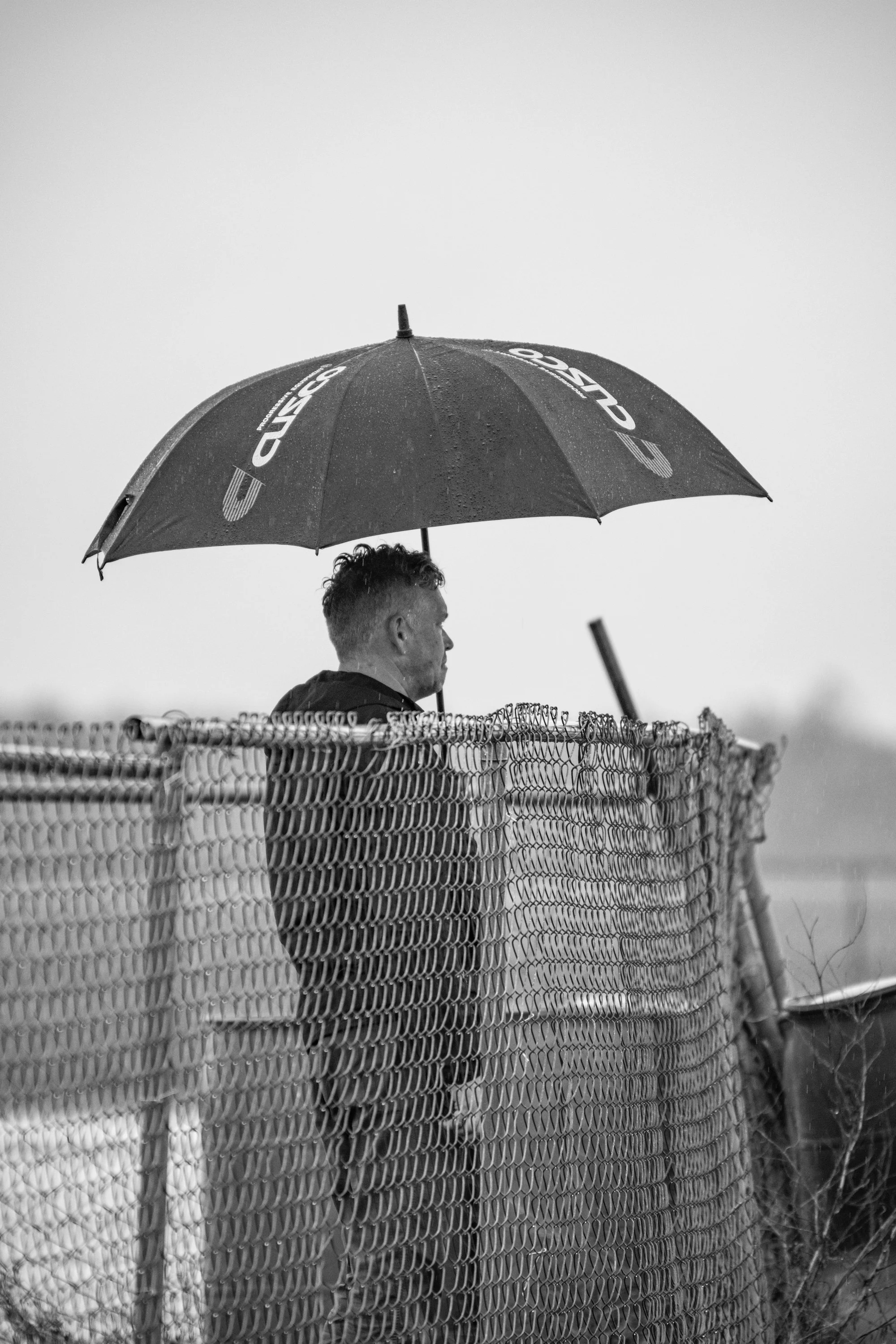 A man standing behind a chain-link fence, holding a large umbrella, in an outdoor setting on a rainy day.