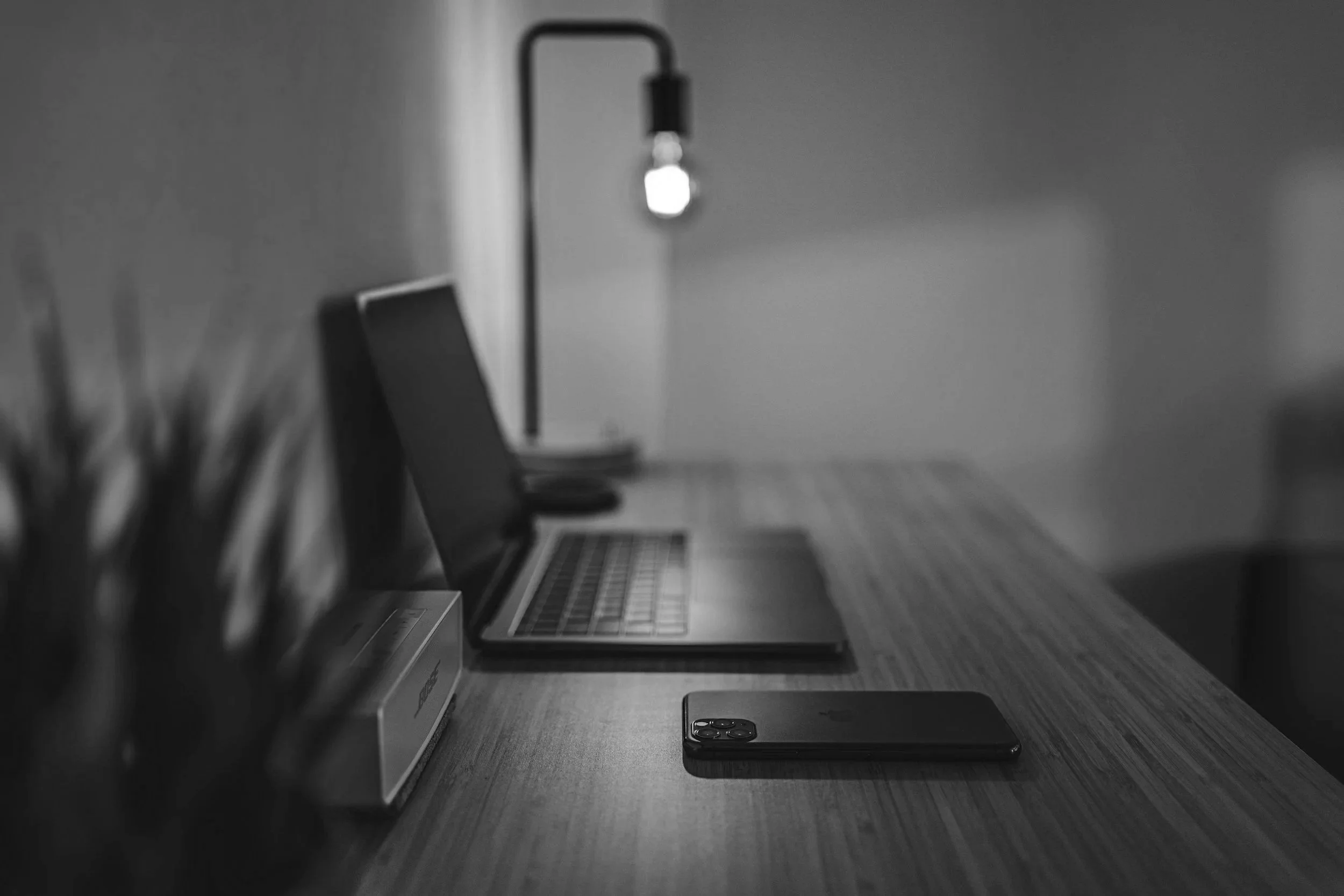 Black and white photo of a work desk with a laptop, smartphone, desk lamp, notebook, pen holder, and a small stack of books, in a minimalist style.