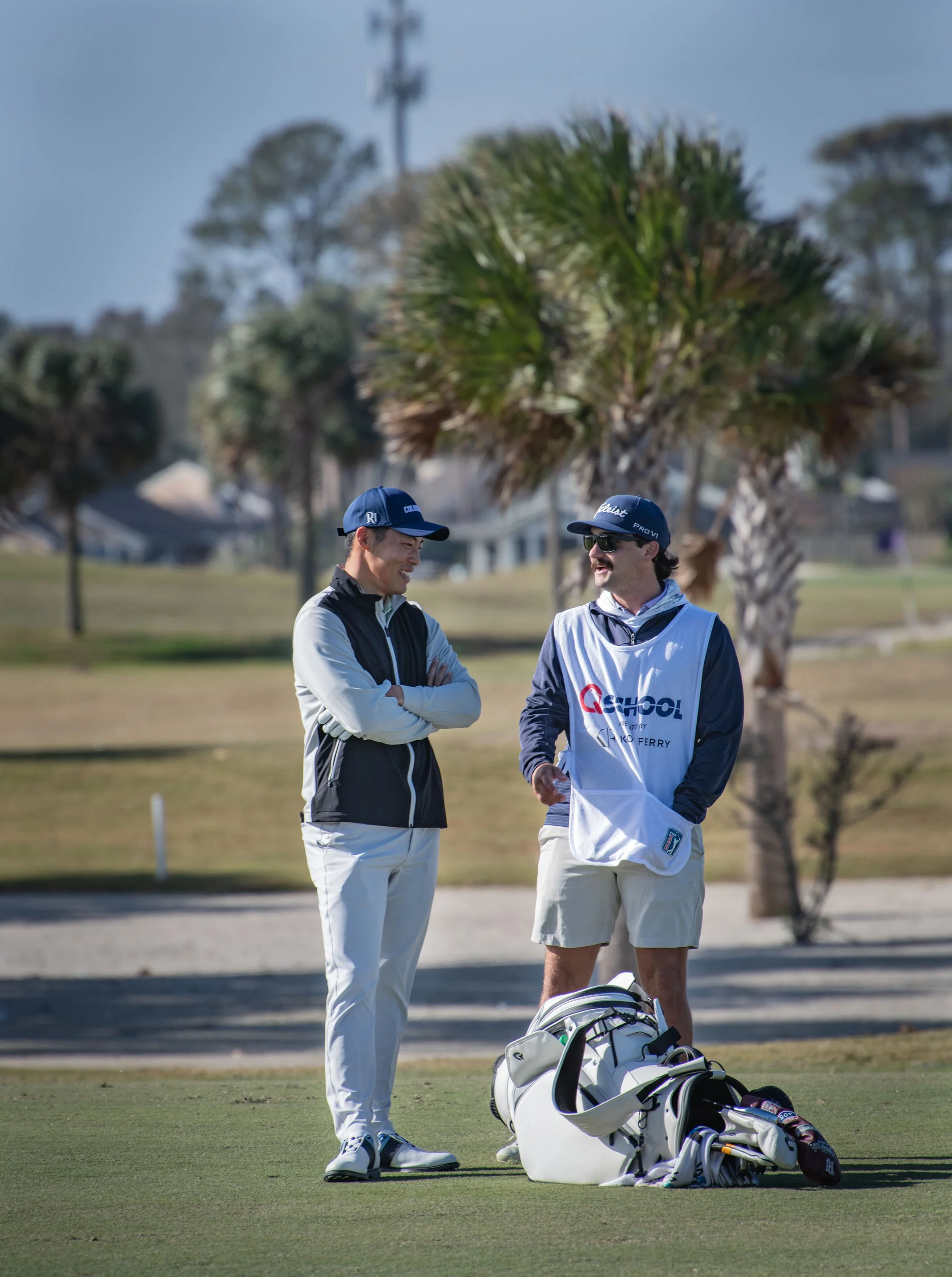 Two golfers on a golf course, one with arms crossed and the other holding a golf tee, standing near golf bags and equipment, with trees and houses in the background.