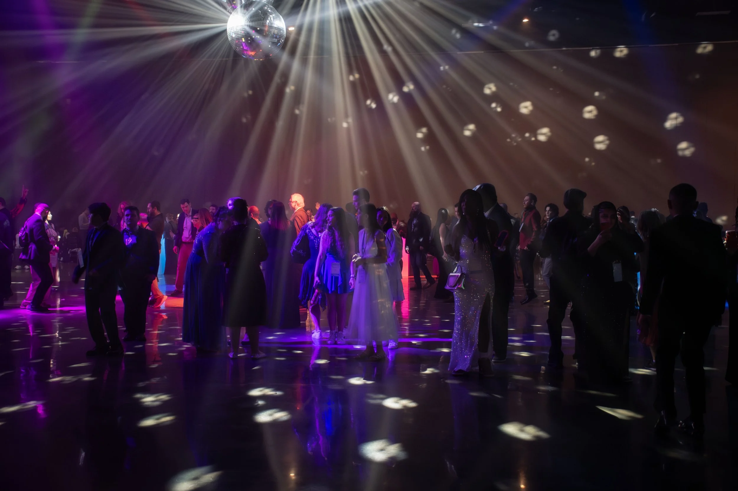 People dancing in a dark, brightly lit dance floor with a mirror ball and colorful light effects.