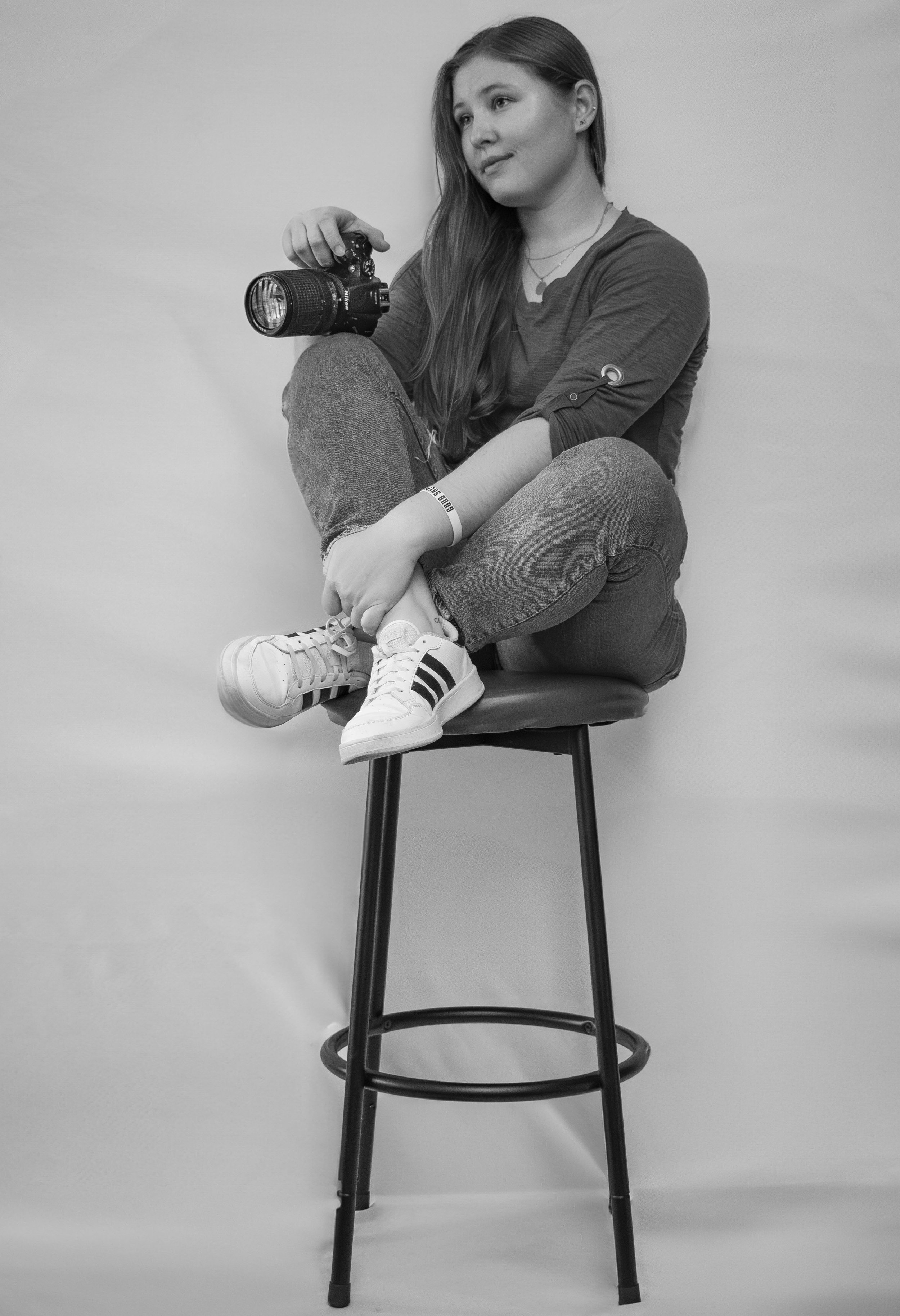 A young woman with long hair and casual clothing sitting cross-legged on a tall stool, holding a camera, against a plain background.