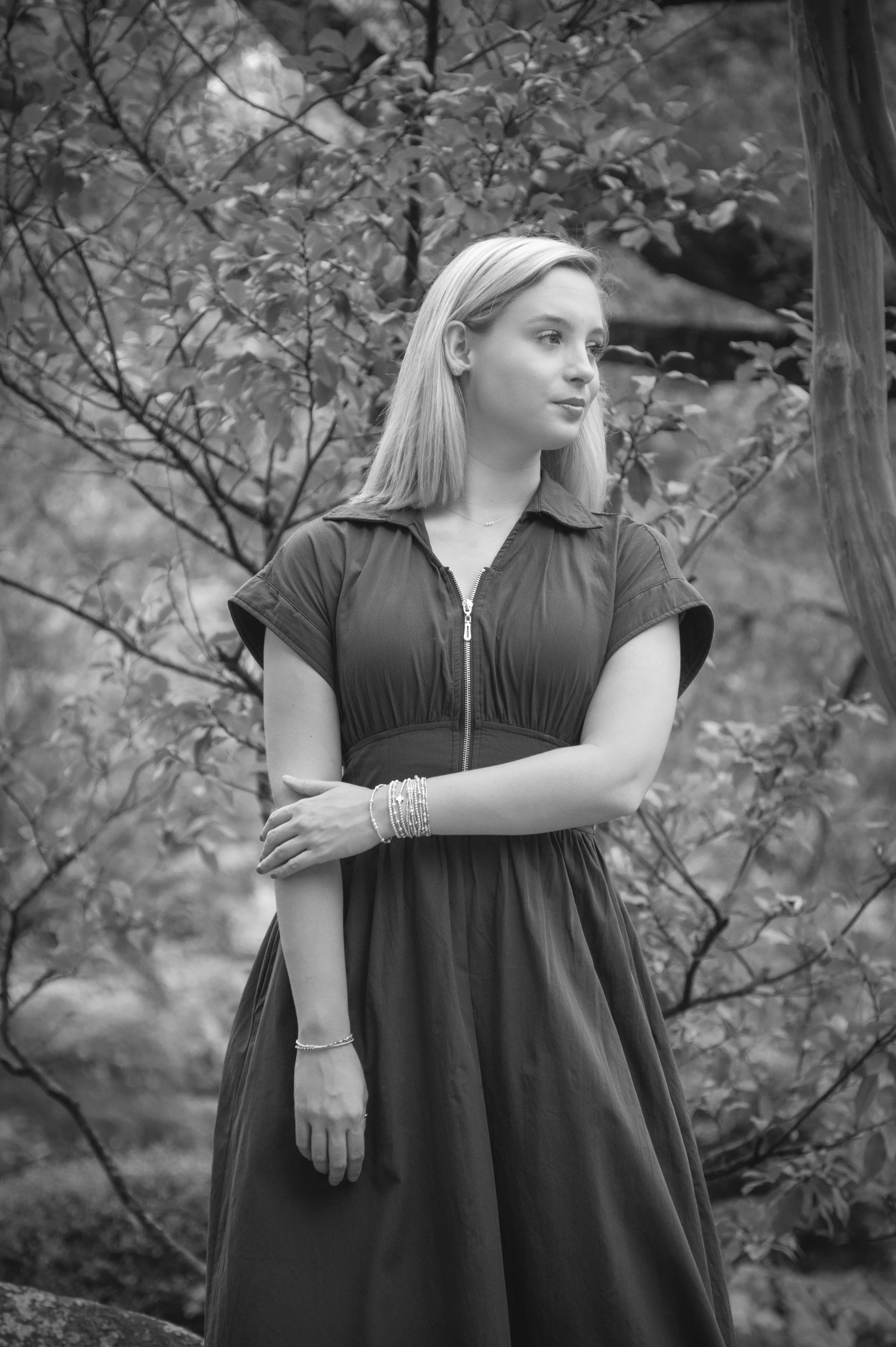 Black and white photo of a young woman with blonde hair standing outdoors, surrounded by trees and foliage, looking to her right with arms crossed.