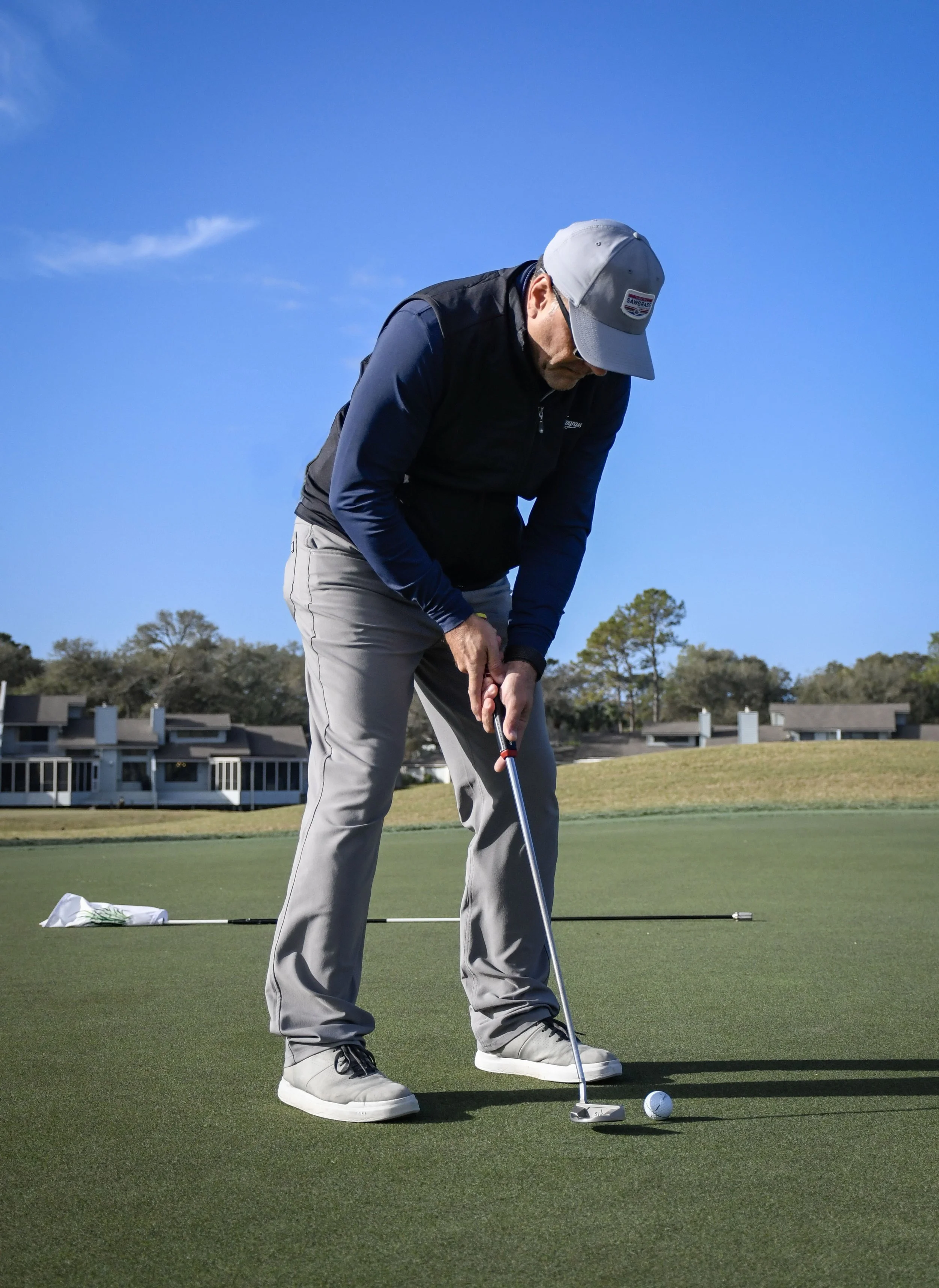 A man is standing on a golf course preparing to putt a golf ball on the green, wearing a gray cap, dark vest, and light pants, with a clear blue sky and trees in the background.