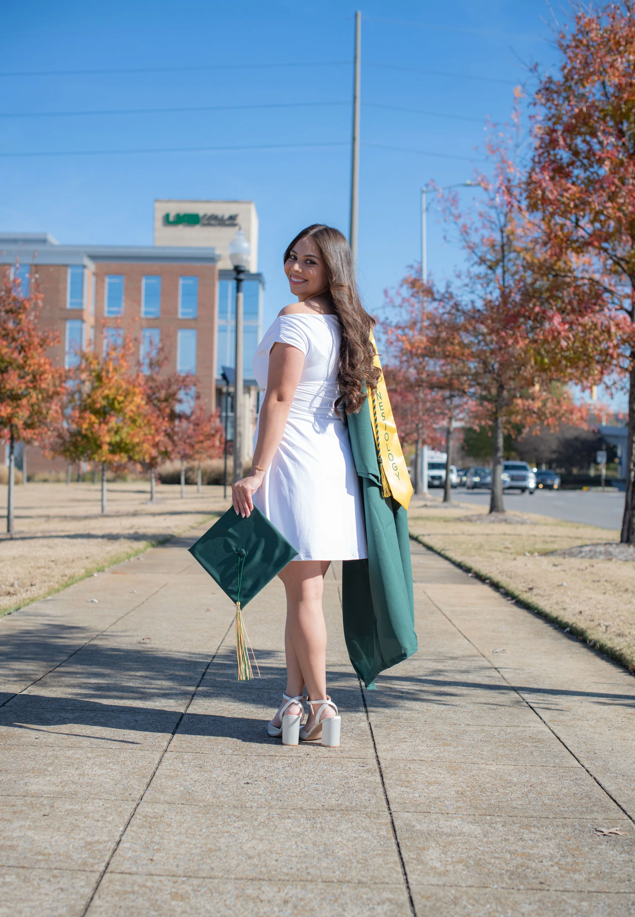 A young woman in a white dress standing outdoors on a sunny day, holding a graduation cap, with a green graduation gown draped over her shoulder, on a sidewalk with autumn trees and a building in the background.