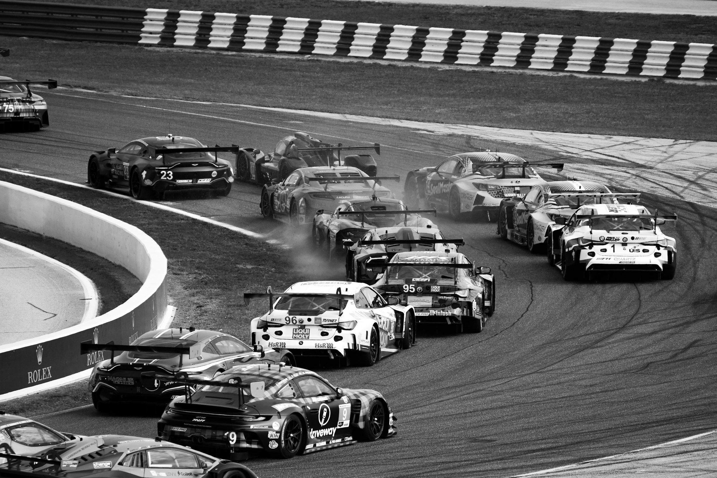 Multiple race cars competing on a track in black and white, some cars are on a curve, leaving tire marks, with a striped barrier at the edge of the track.