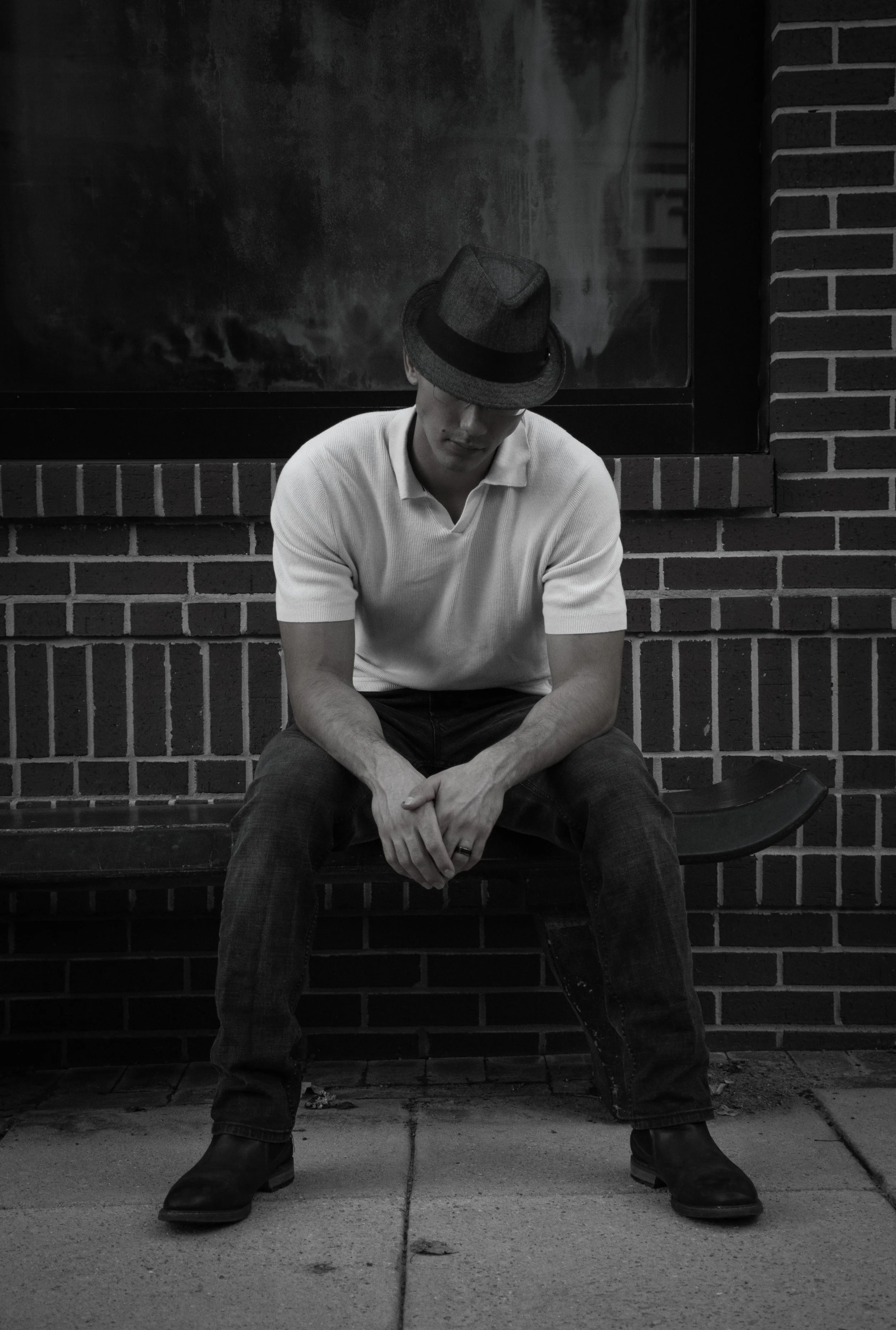 A man sitting on a bench against a brick wall, wearing a fedora hat, a white polo shirt, dark jeans, and boots, with his head bowed and hands clasped.