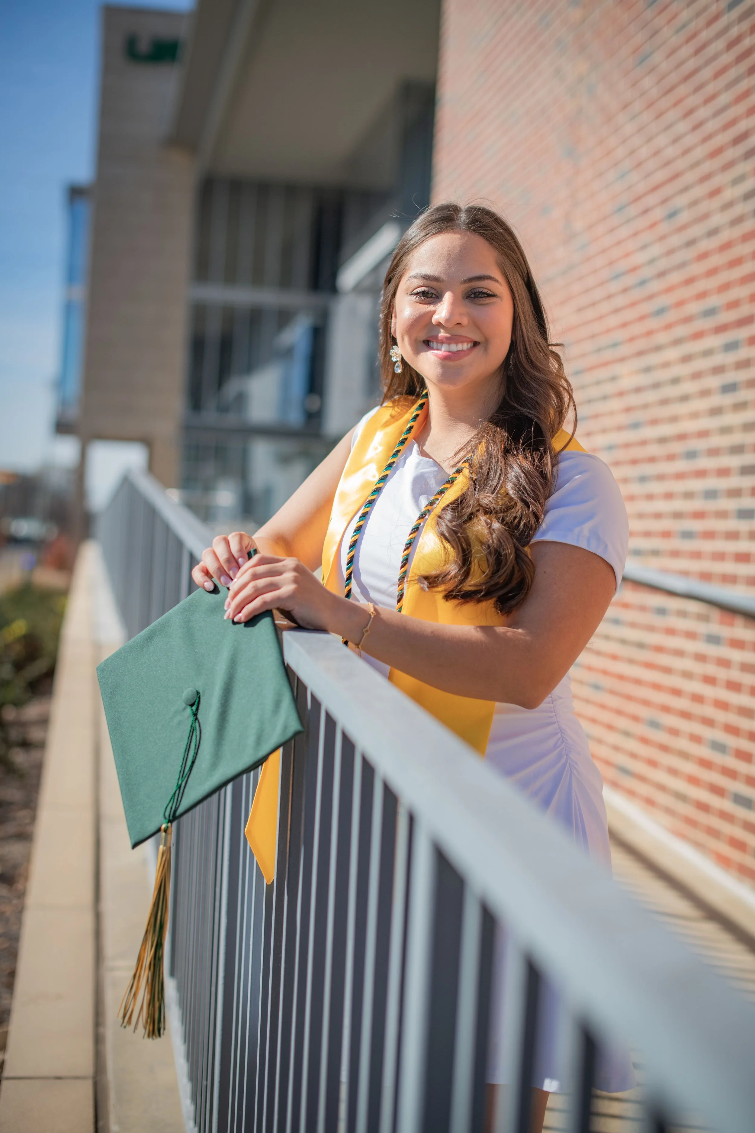 A young woman in a white dress and yellow stole, celebrating her graduation outdoors on a sunny day, holding a green graduation cap.