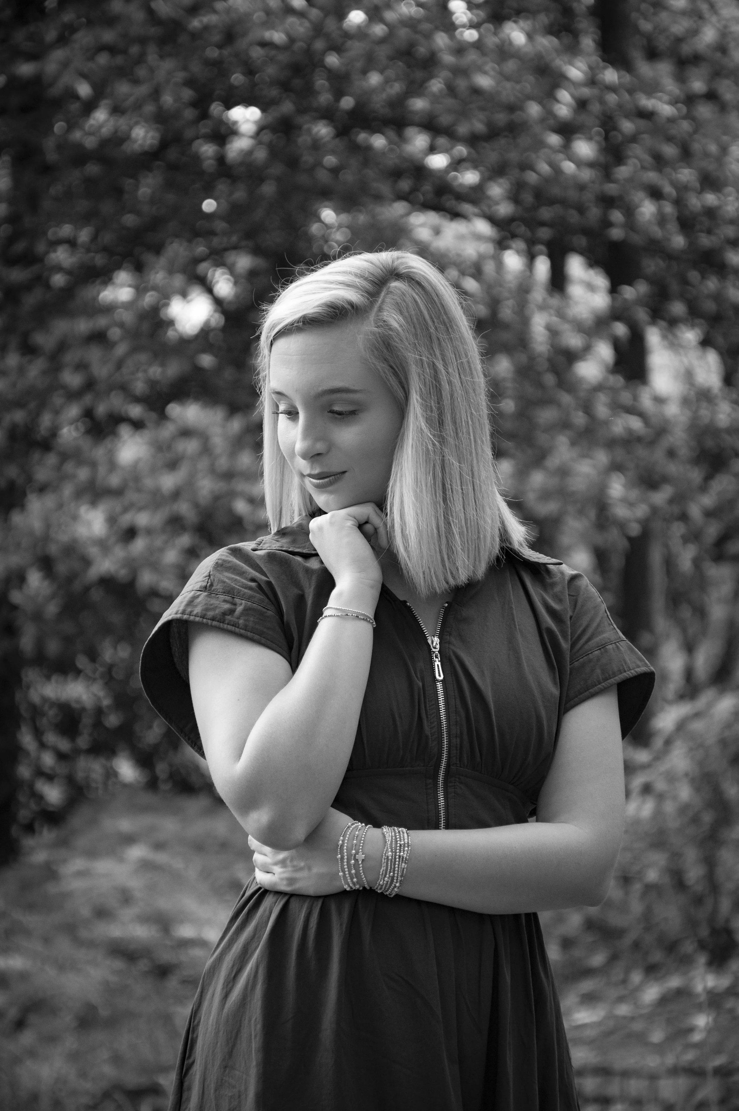 A young woman with shoulder-length hair, wearing a short-sleeved dress with a zipper front, standing outdoors among trees. She is looking down with her hand near her chin, and wearing multiple bracelets on her wrist.