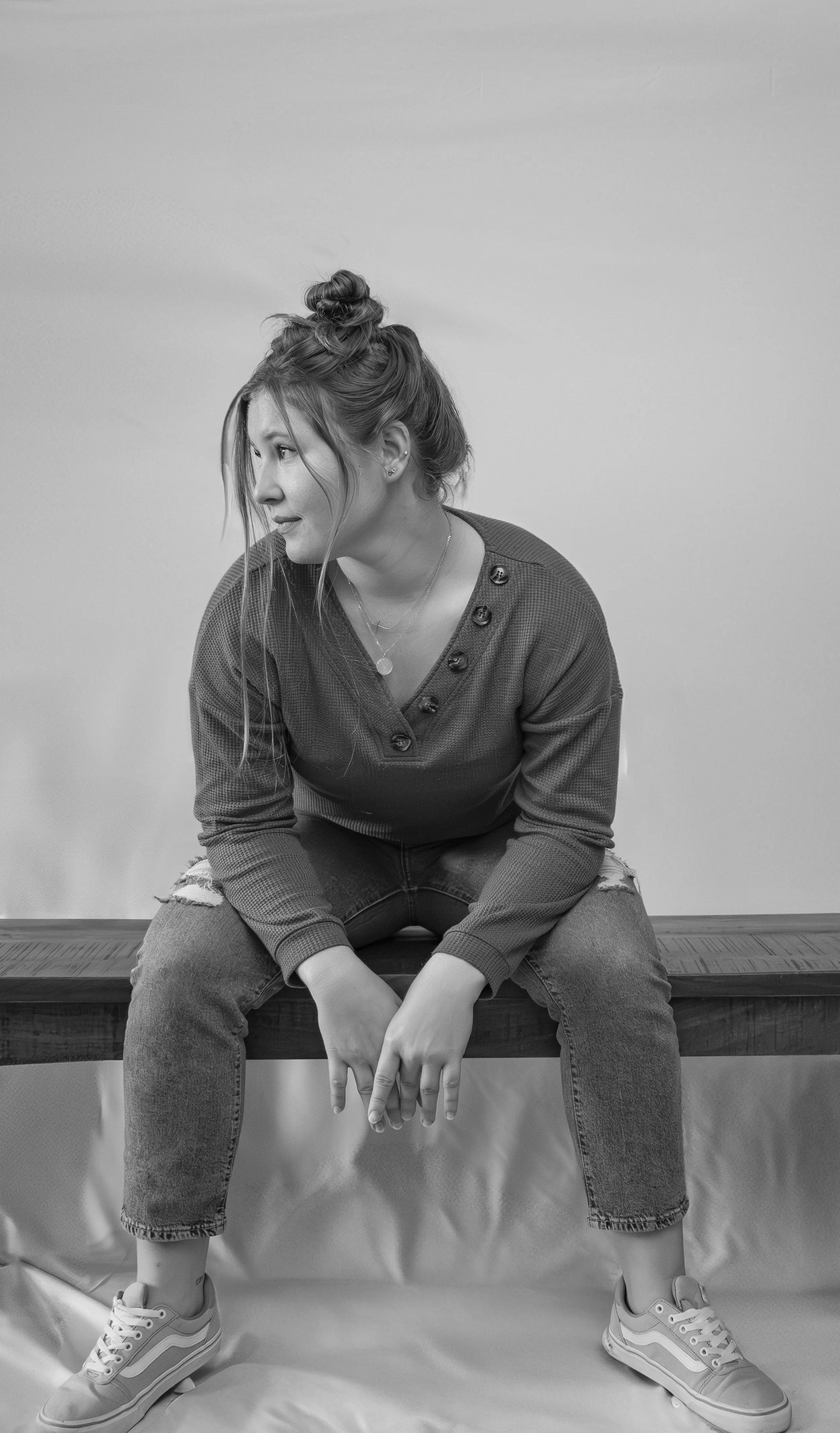 A woman with a casual style, wearing ripped jeans and sneakers, sitting on a wooden bench against a plain wall, looking to the side.