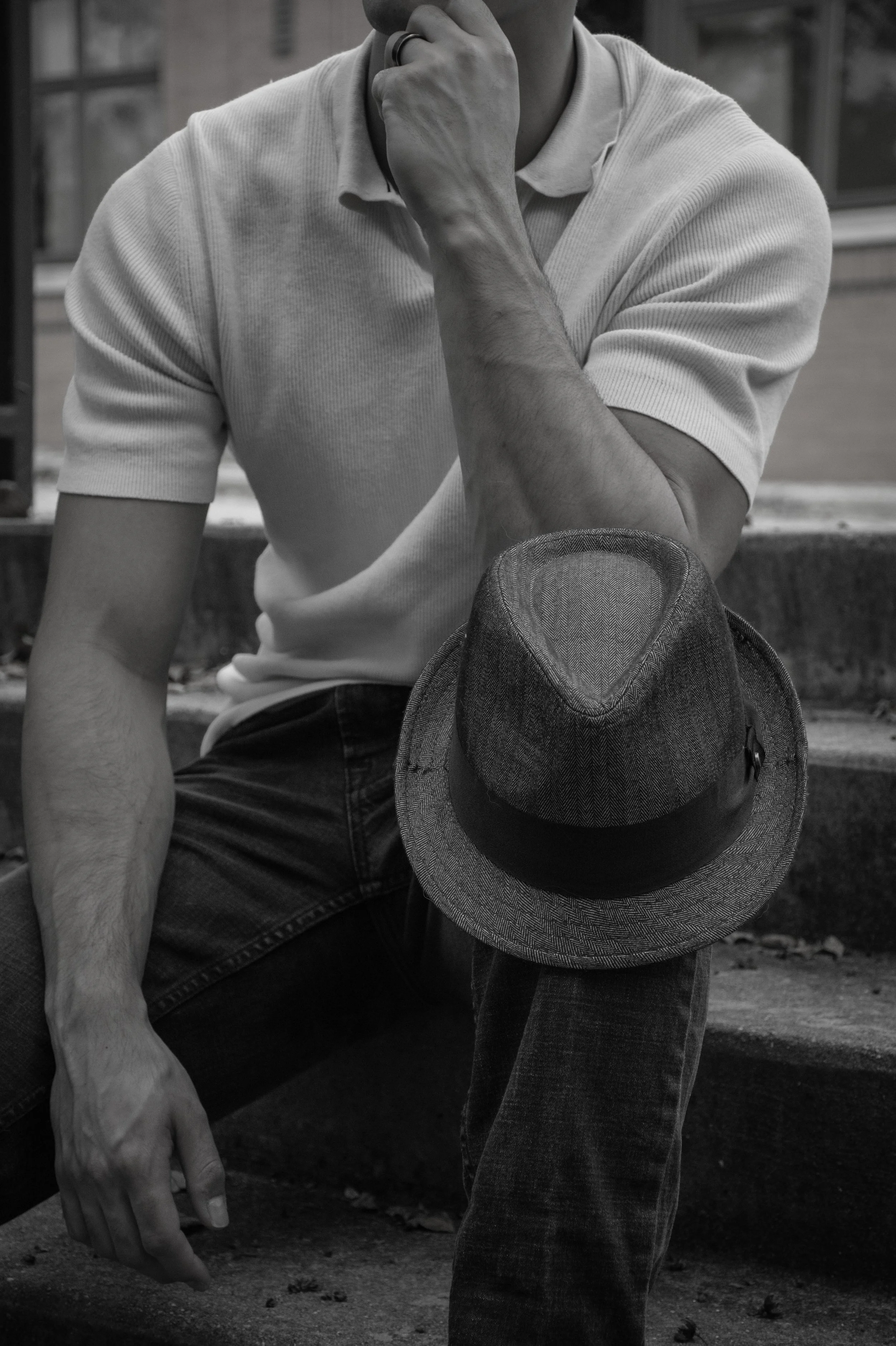 A person sitting on outdoor stairs, wearing a light-colored shirt and dark jeans, resting with a fedora hat hanging from their knee, black and white photo.