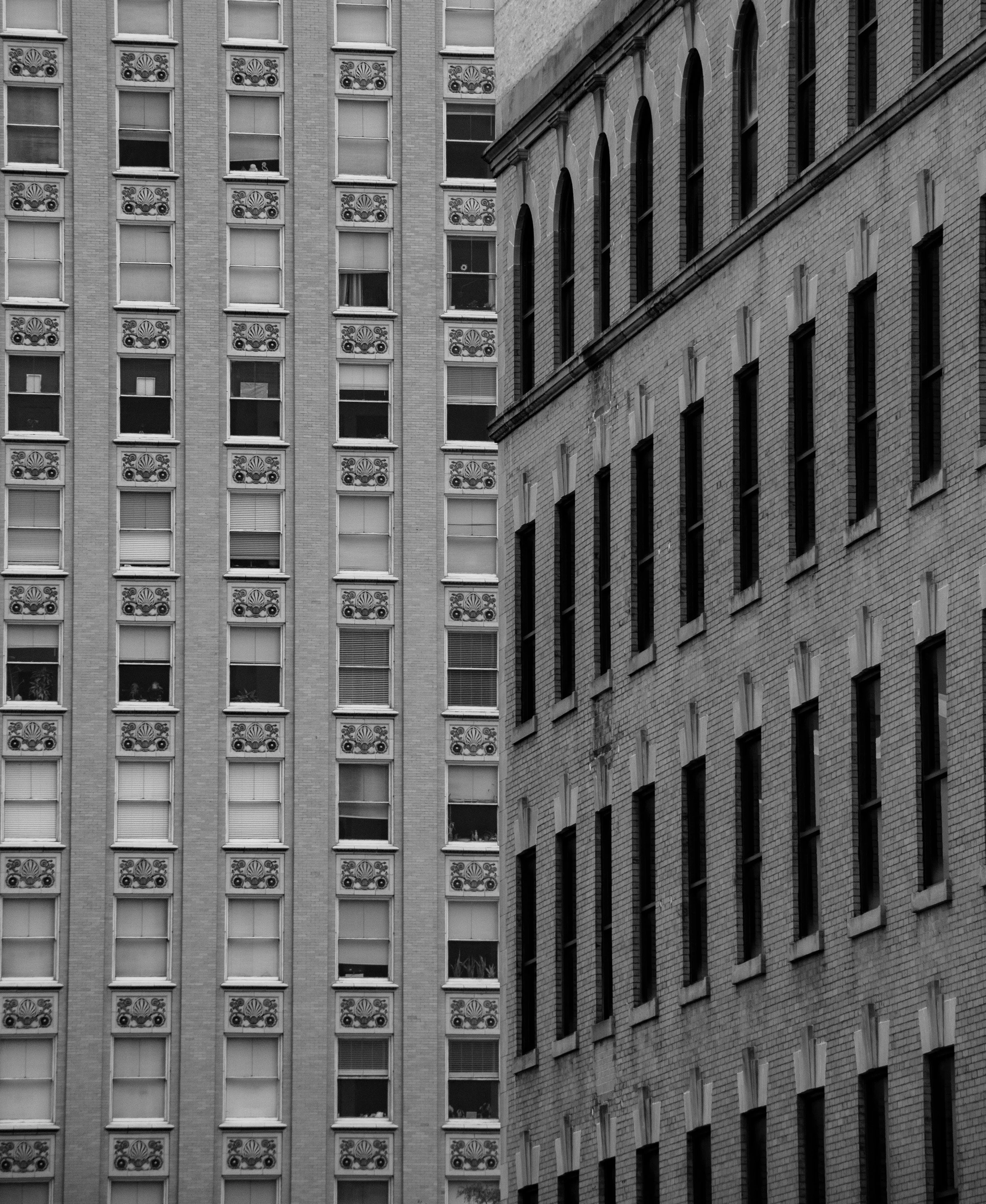 Black and white photo of tall buildings in an urban city, with the building on the left featuring decorative floral patterns on the window frames and the building on the right showing a brick facade with arched and rectangular windows.