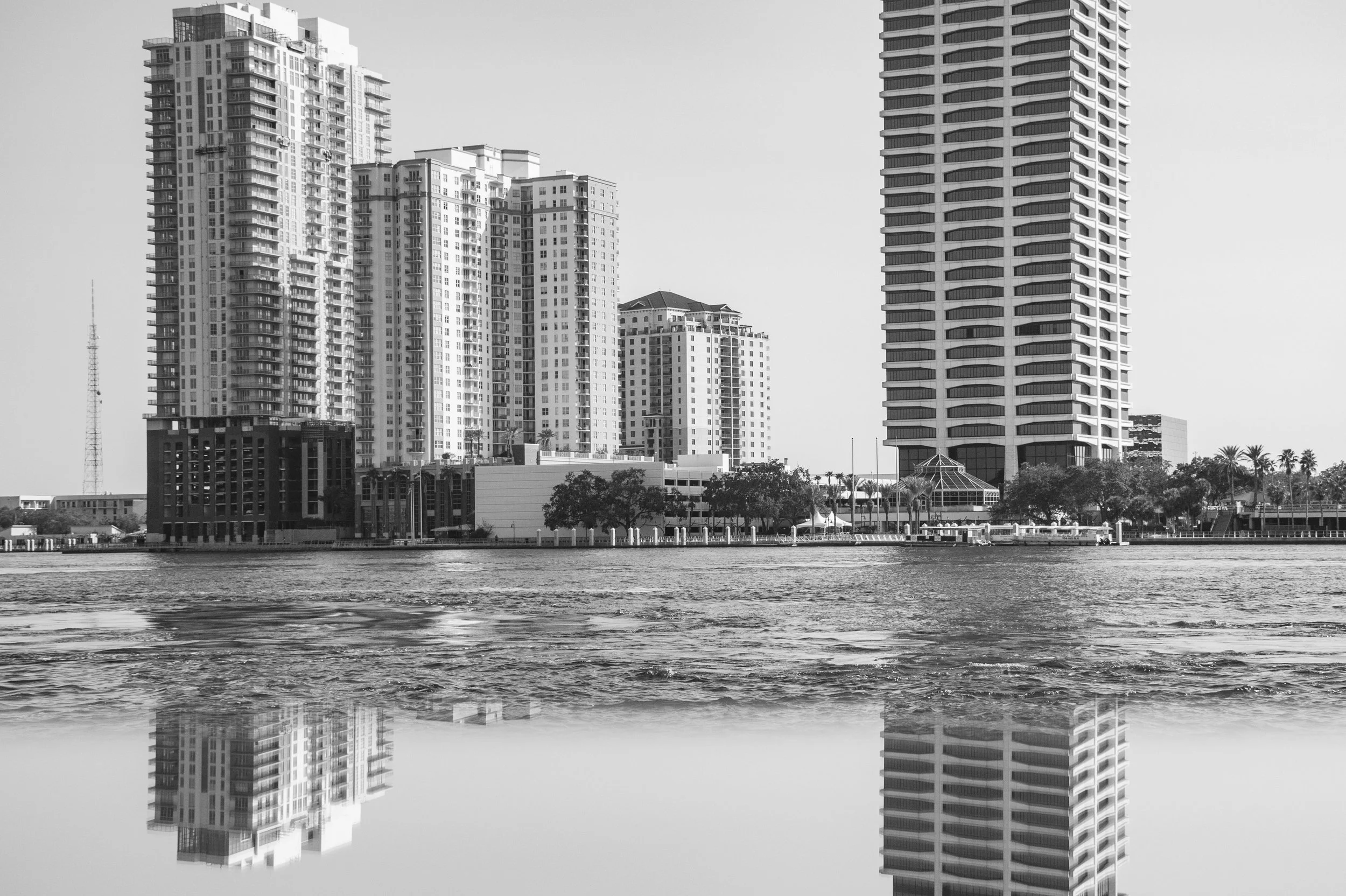 Black and white photo of tall modern high-rise buildings by a body of water, with reflections visible in the water.