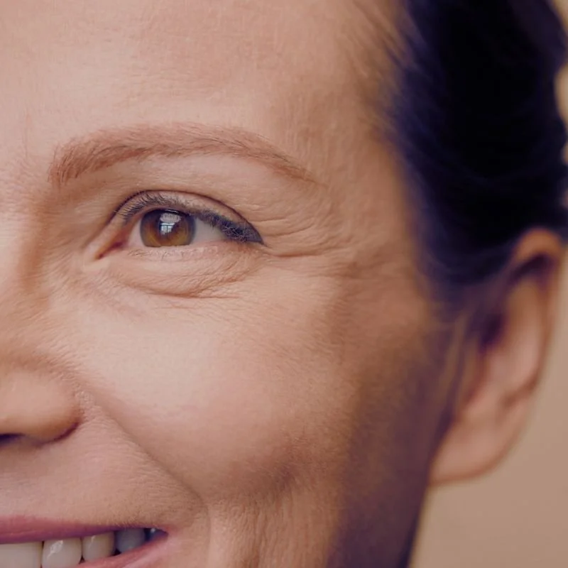 Close-up of a smiling woman's face showing her brown eye, cheek, and part of her nose with a smooth complexion.