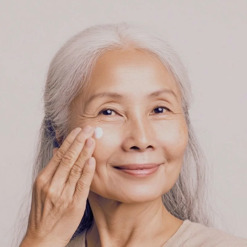 An elderly woman with long gray hair applying moisturizer to her face with her right hand and smiling.