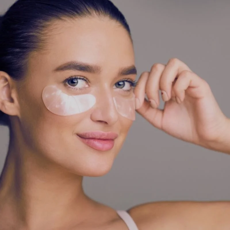 A woman with dark hair slicked back, applying under-eye patches, looking at the camera with a slight smile.