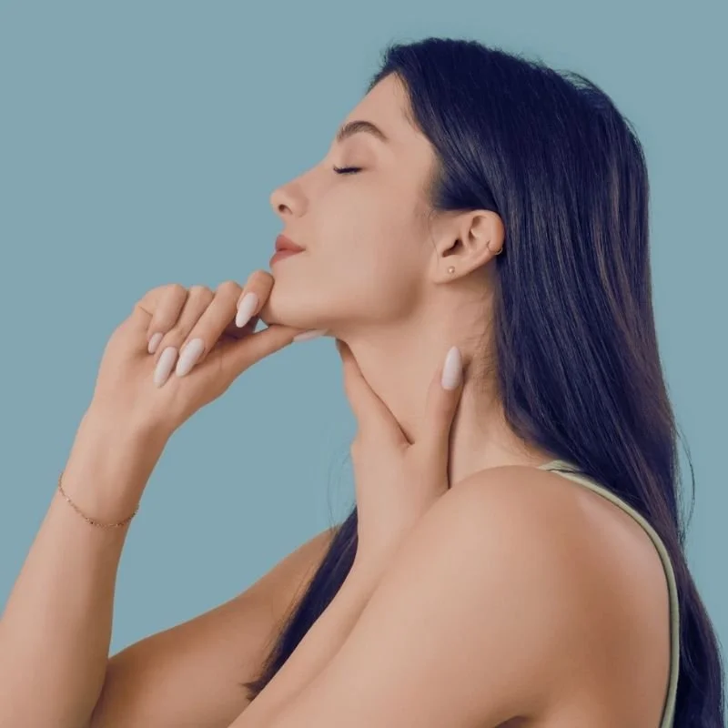 A young woman with long dark hair and light skin poses with her eyes closed, touching her chin and neck with both hands, against a blue background.
