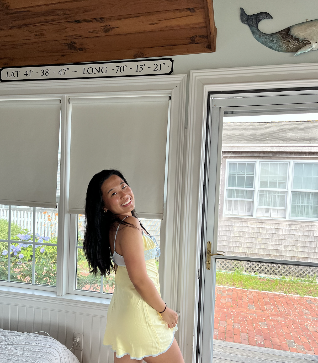 A young woman in a floral dress standing in front of a window with a floral arrangement on the side.