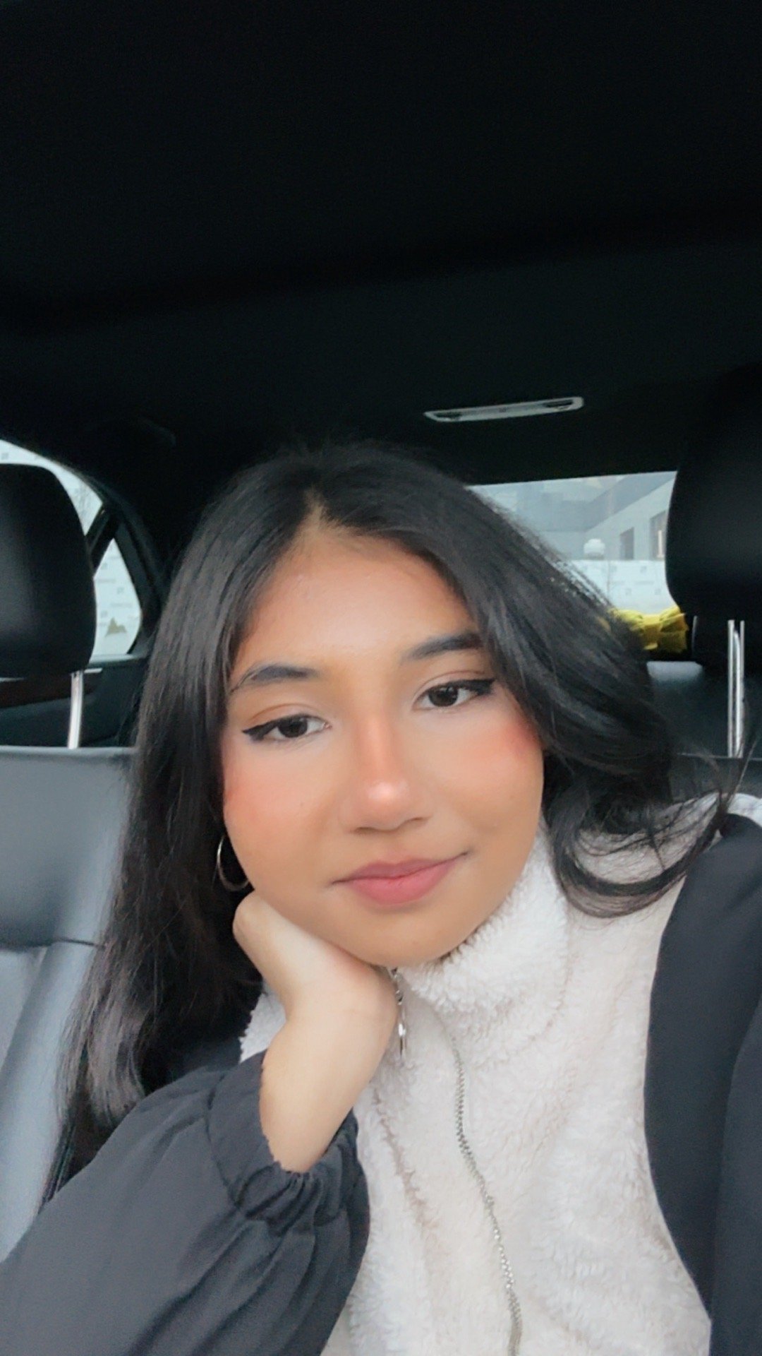 A young woman with long brown hair and a white smile, wearing a light gray sweatshirt with university lettering, jeans, and white sneakers, standing outdoors on a curved pathway covered with fallen autumn leaves, in a park with trees, benches, and a black lamp post, during daytime.