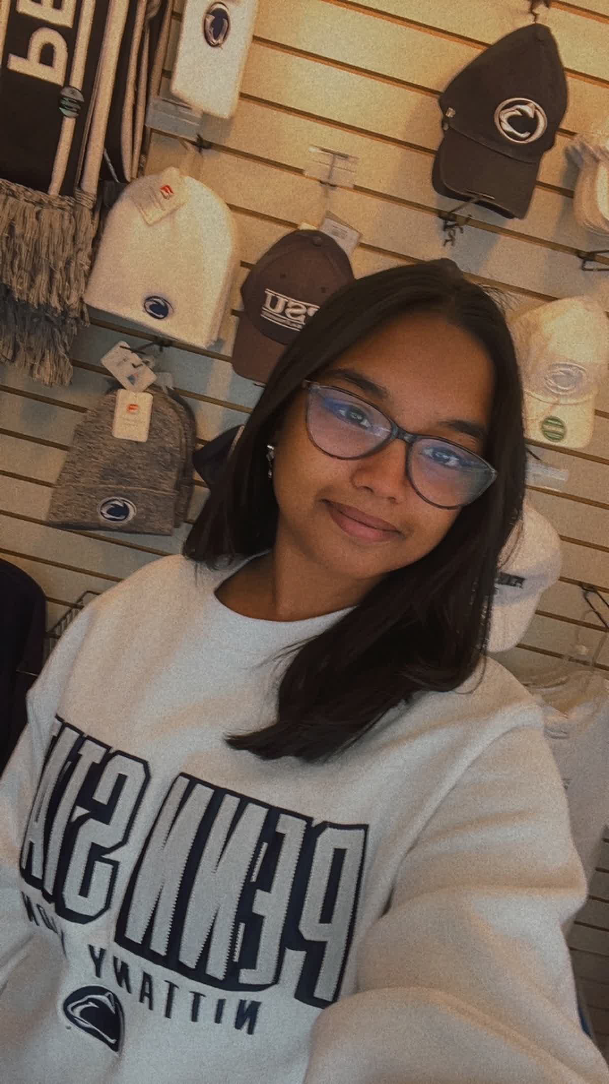 Woman wearing glasses in a turquoise saree sitting on wooden steps, holding a flute, with large potted plants in the background.