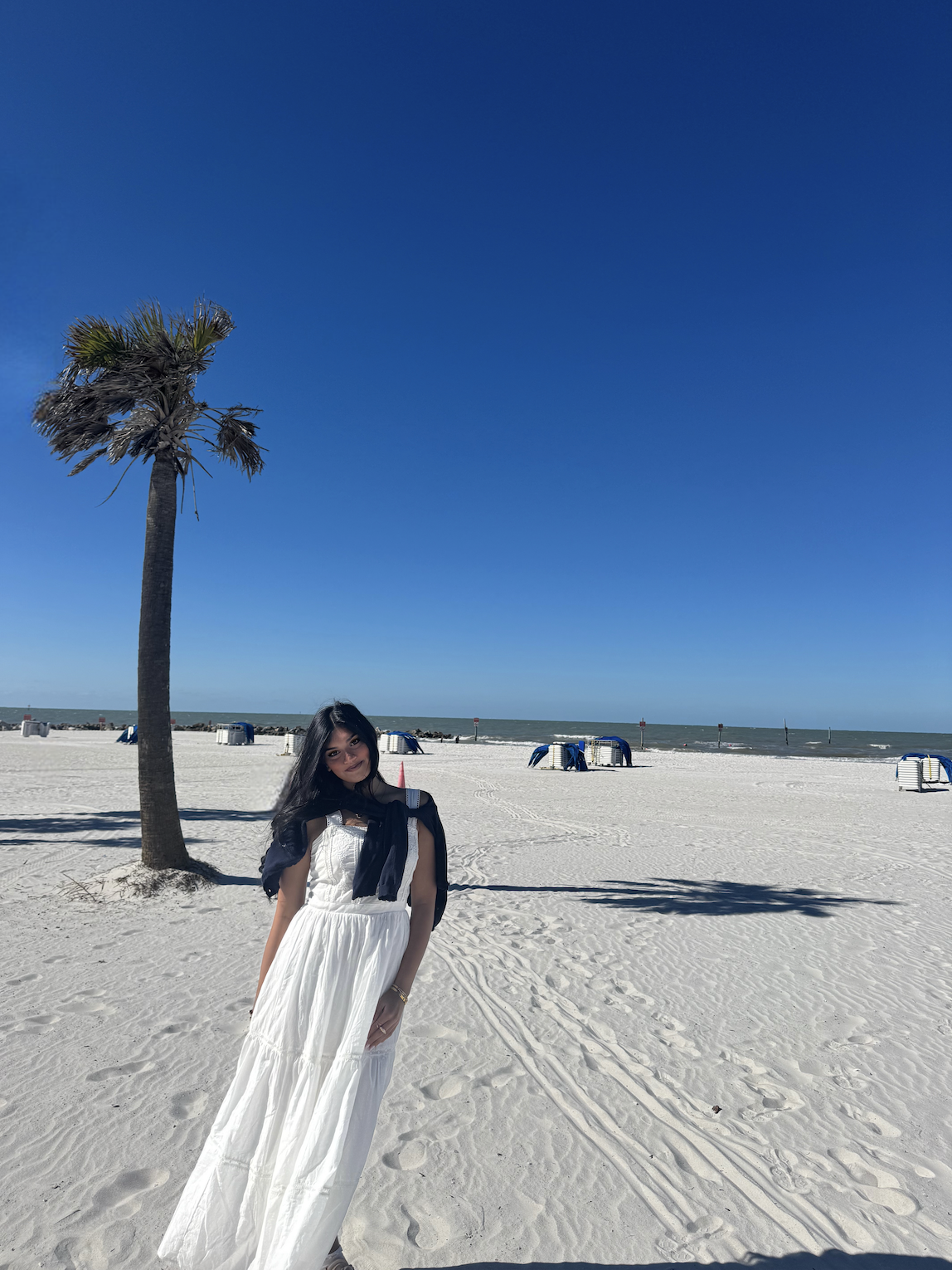 A woman sitting on a wooden bench under a large tree, smiling at the camera in an outdoor setting with green grass and trees in the background during daylight.