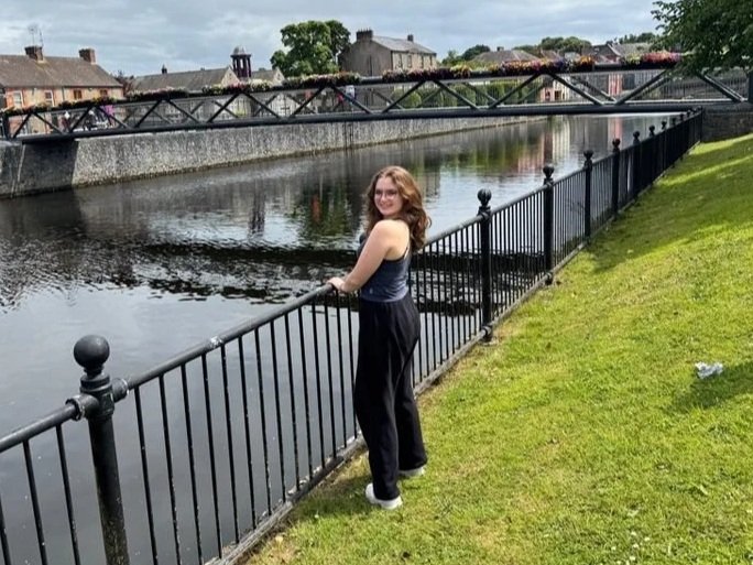 Young man smiling outdoors at a soccer field during sunset, wearing a Penn State Men's Soccer t-shirt with a hoodie underneath.
