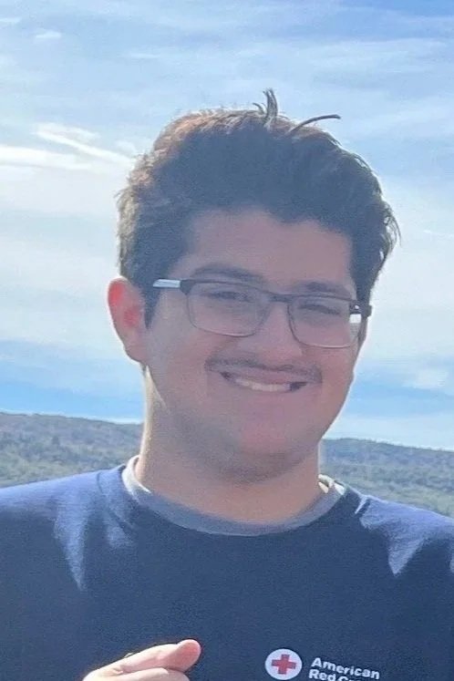 A young man with black hair, glasses, and a badge around his neck taking a selfie in a busy indoor setting.