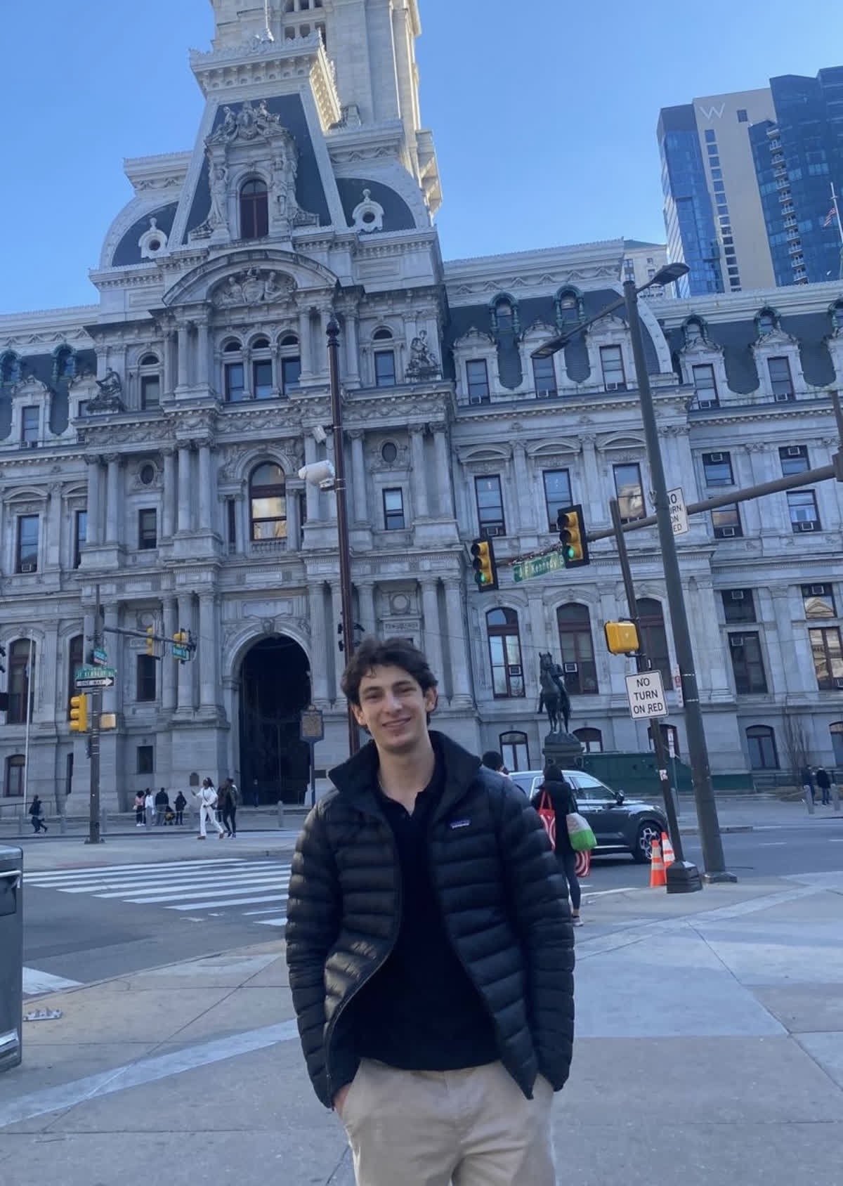 A woman with long black hair, glasses, and jewelry stands outdoors under a covered structure, with a park and greenery in the background.