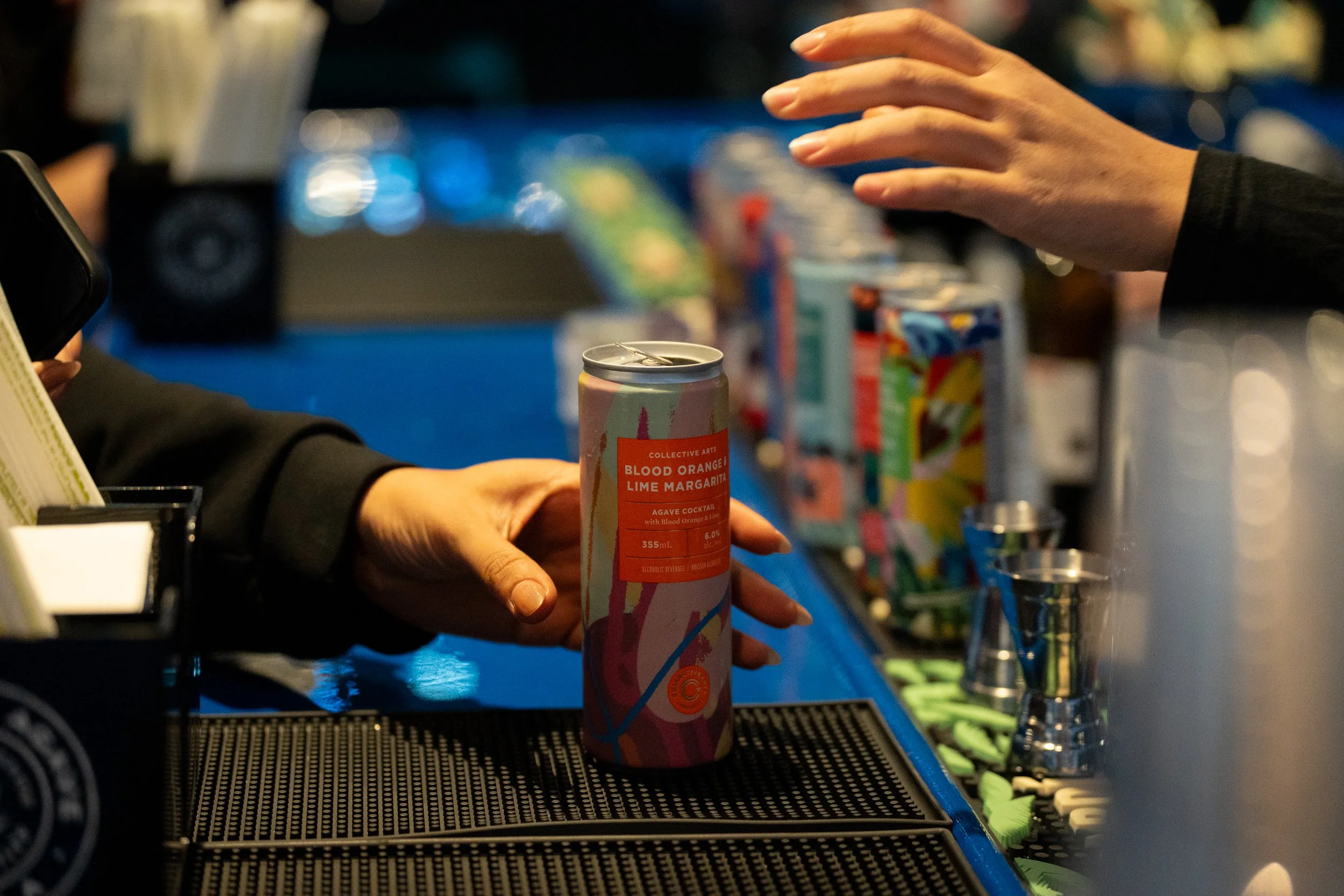 A person's hand reaching to take a can of Blood Orange Lime Margarita from a bar counter. The bartender's hand is visible, and colorful drink cans are blurred in the background.