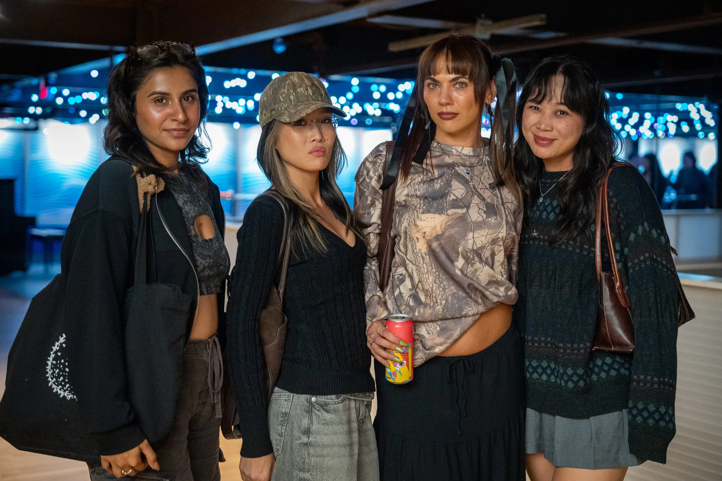 Four women standing in a bar or club with blue string lights, wearing casual clothes, posing for the photo.