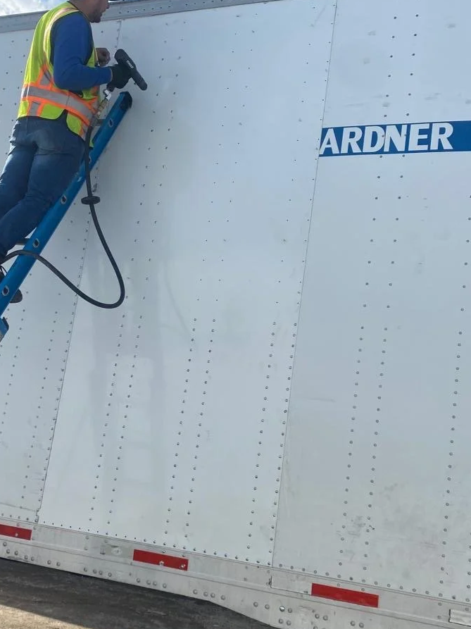 Worker in a safety vest cleaning or inspecting a large white trailer wall with a power tool, partially visible ladder, and a blue company logo that reads 'ARDNER'.