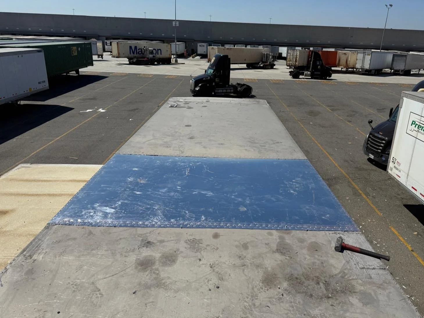 View from above of a loading dock with trucks and trailers parked, showing a freshly installed blue and grey reflective tape on the concrete surface, and a hammer resting on the ground.