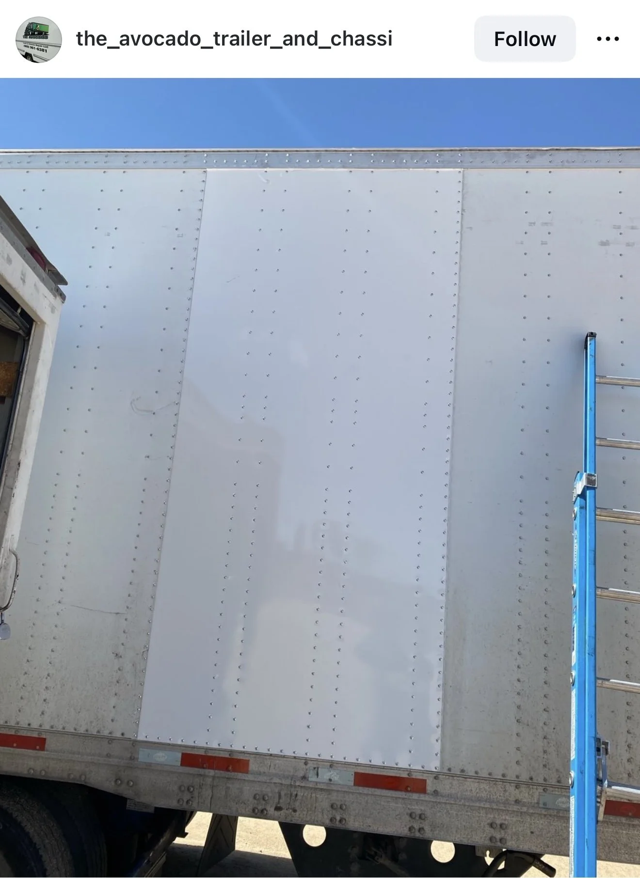 The side of a white trailer with rivets, showing part of a blue ladder and the rear door of the truck.
