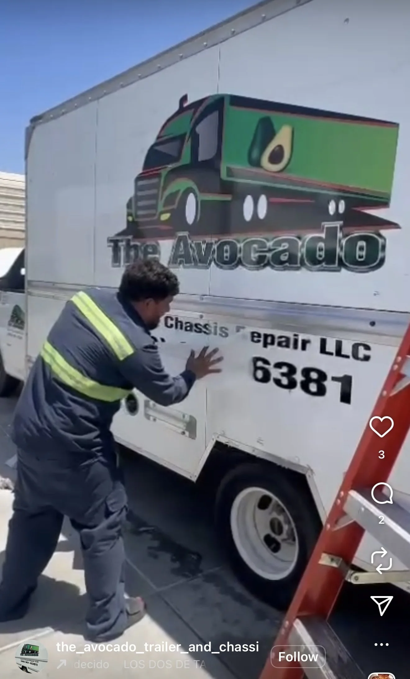A worker attaches a decal to a white trailer truck with the logo of 'The Avocado' company, featuring a cartoon avocado truck, with nearby ladder and sunlight.
