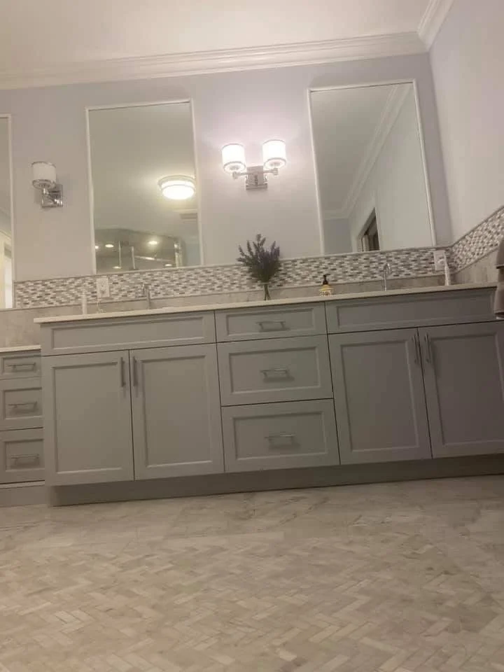 Bathroom vanity with gray cabinets, two mirrors, a small potted plant, and a mosaic tile backsplash.