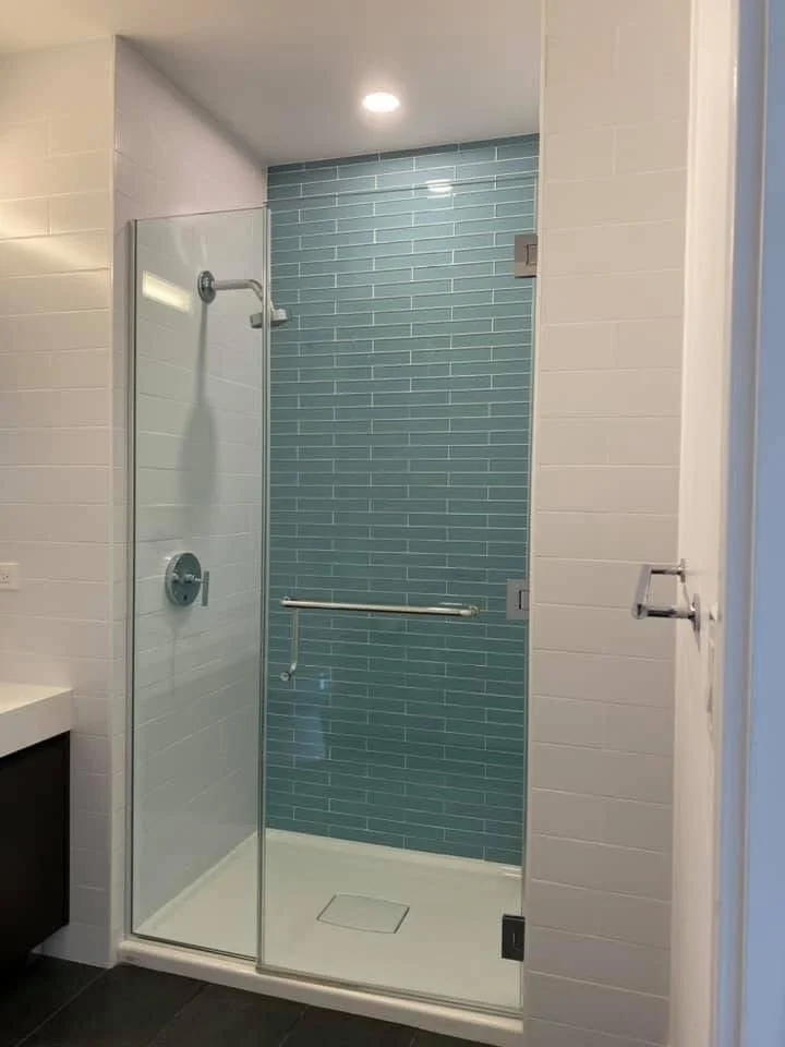A modern walk-in shower with a glass door, blue subway tile wall, a ceiling light, a showerhead, a control valve, and an integrated grab bar.