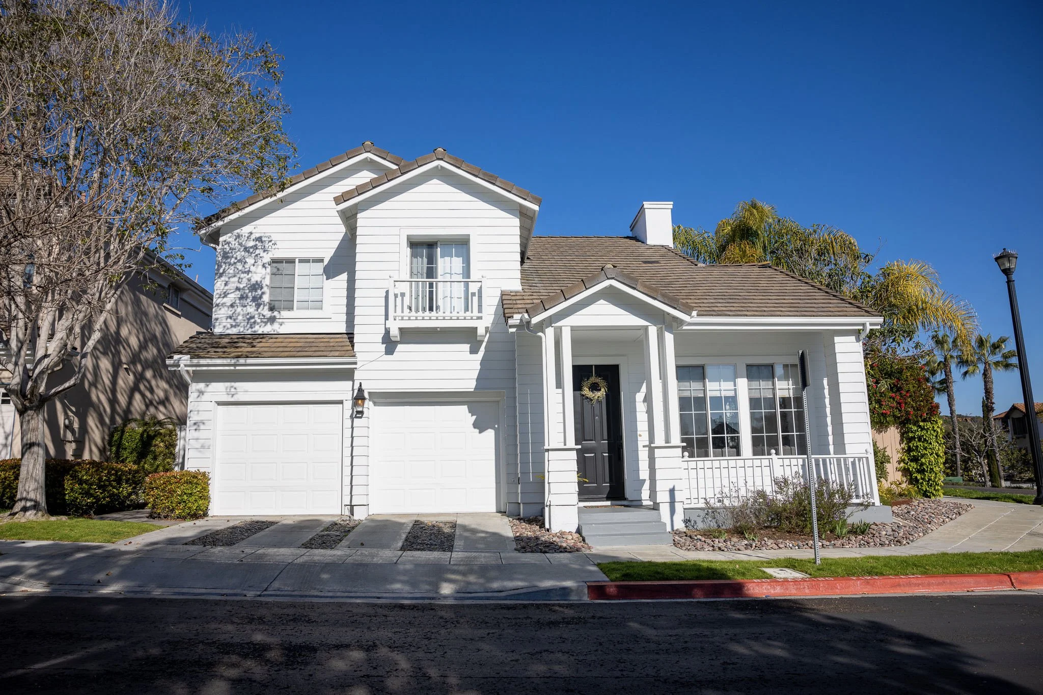Tidewell Properties - Front view of a white two-story house with a small front porch, garage doors, and a streetlamp, under a clear blue sky.