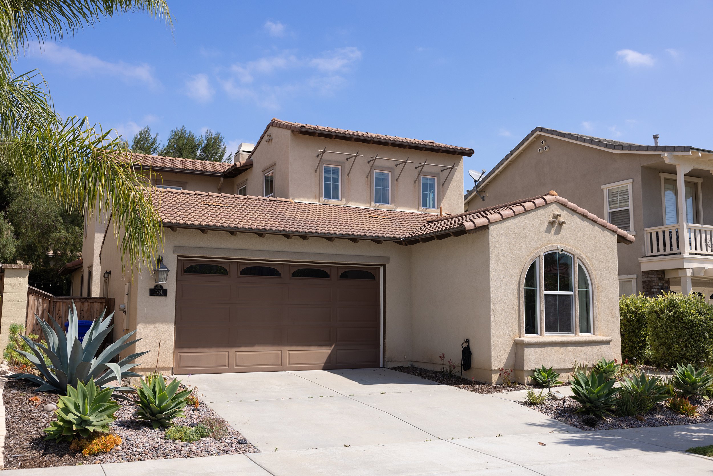 A two-story beige house with a brown garage door, a rounded window, and a front yard with desert plants and succulents. Blue sky with some clouds in the background.
