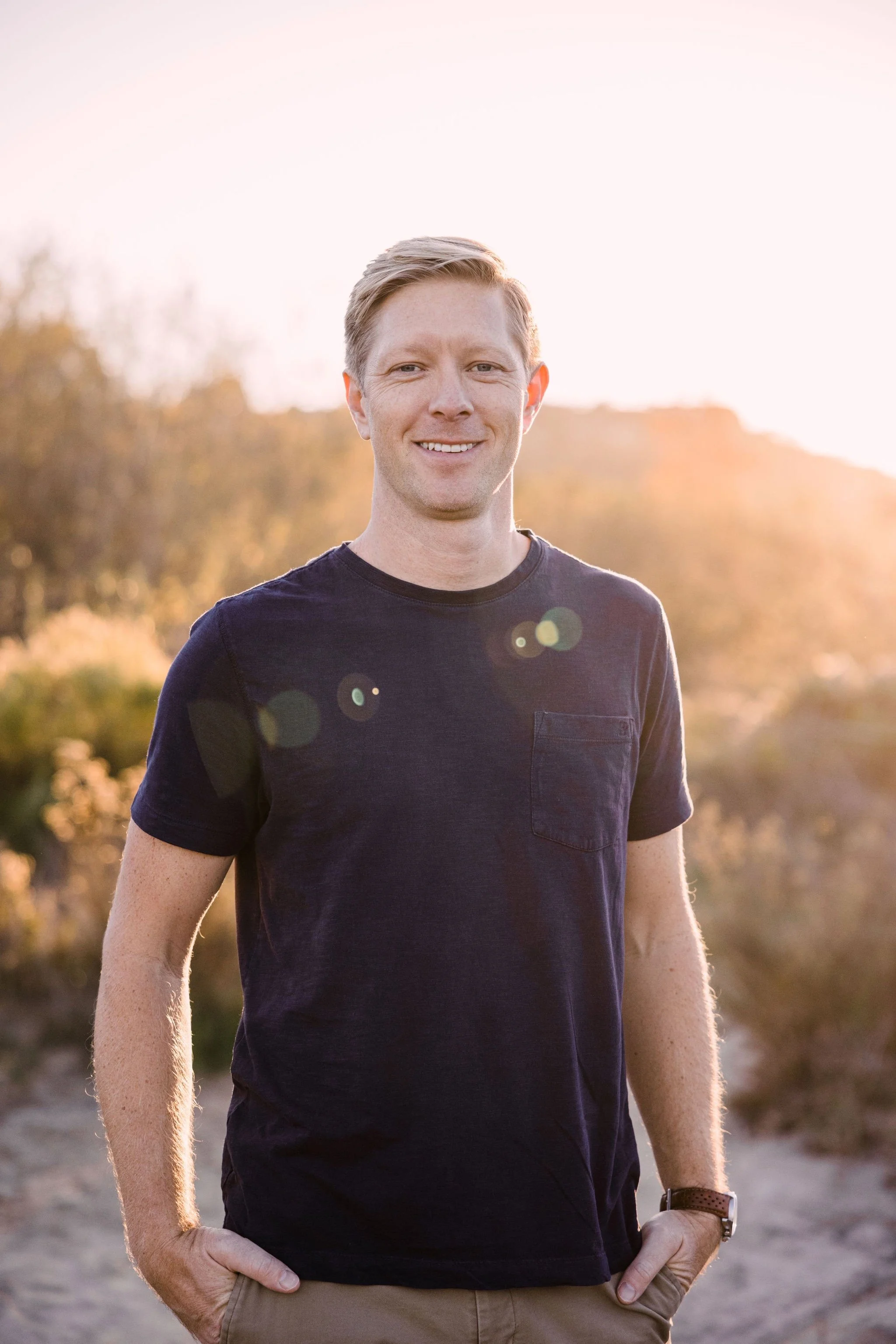 Founder Shane Fellows - standing outdoors during sunset, wearing a navy blue t-shirt, with trees and a hill in the background.