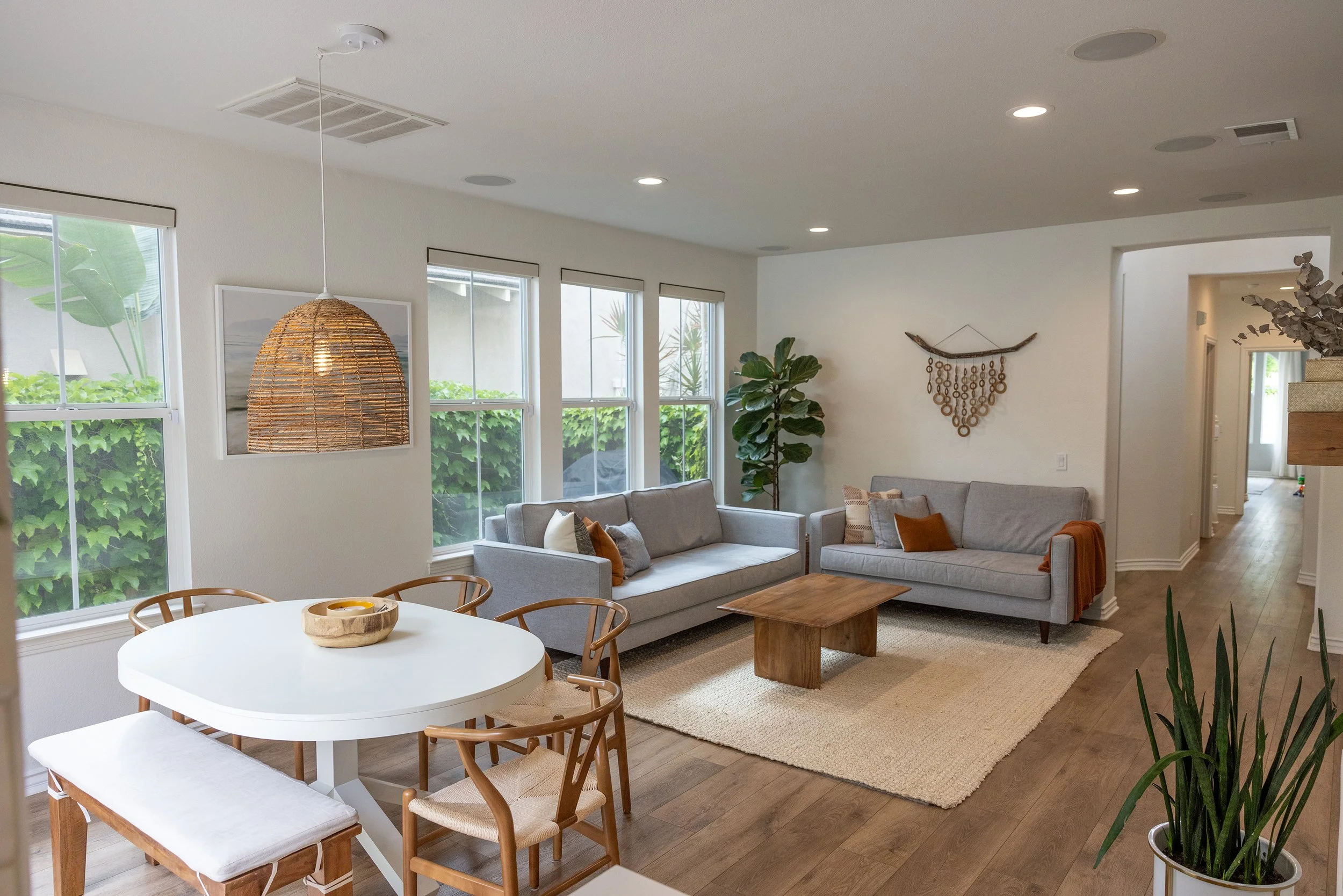 Tidewell Properties - Living room with white walls, large windows, and wooden flooring, featuring two gray sofas, a wooden coffee table, a beige rug, potted plants, wall decorations, and a dining area with a white round table and wooden chairs.