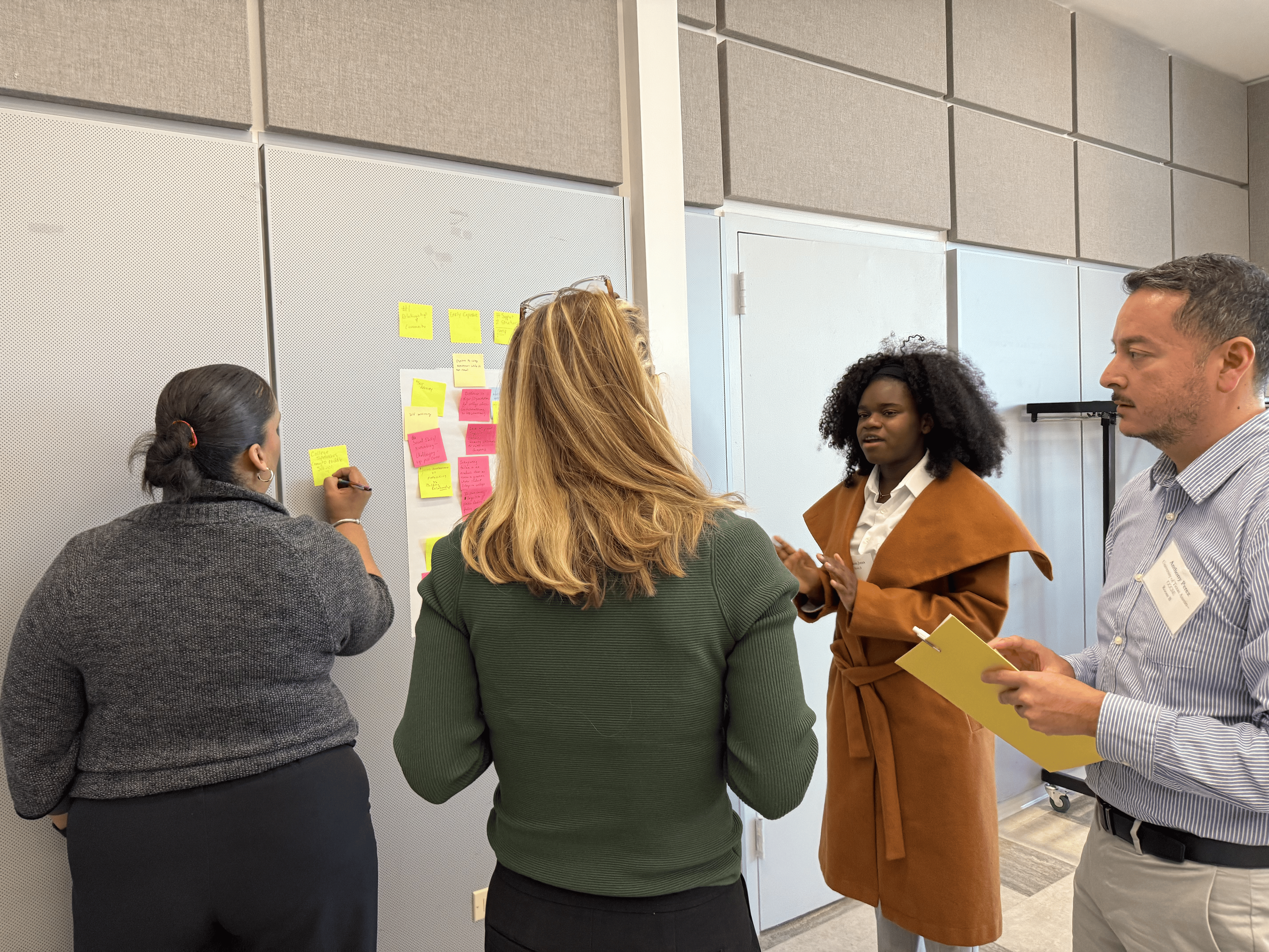 Four people engaged in a discussion around a wall with yellow and pink sticky notes in a conference room.