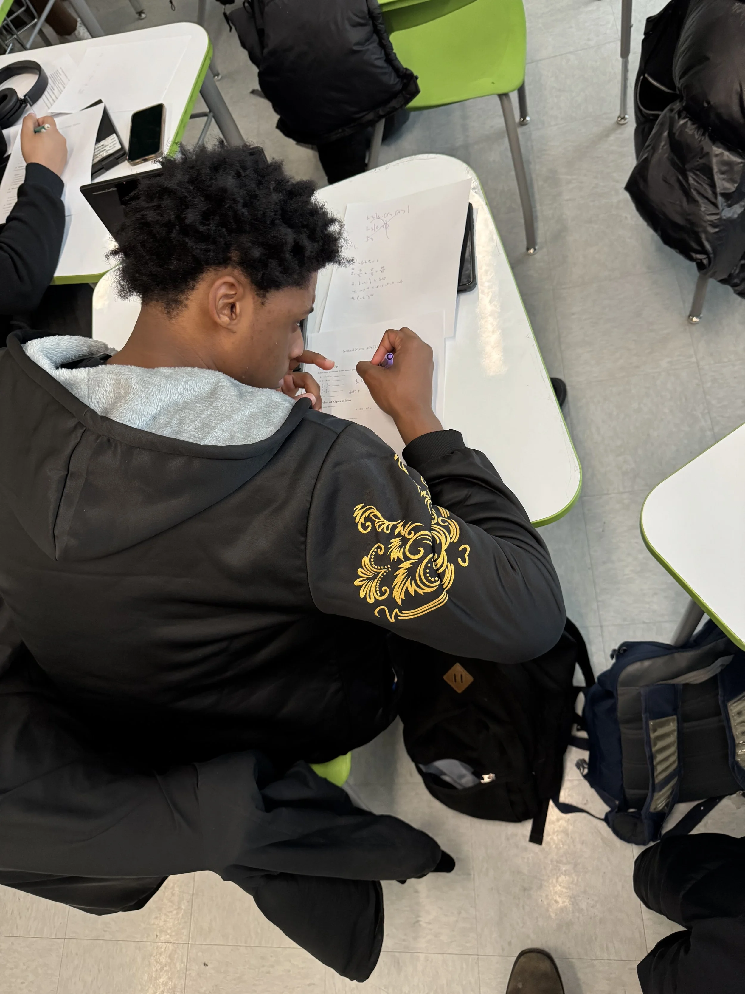 A student sitting at a desk, writing on paper with pens, studying in a classroom.