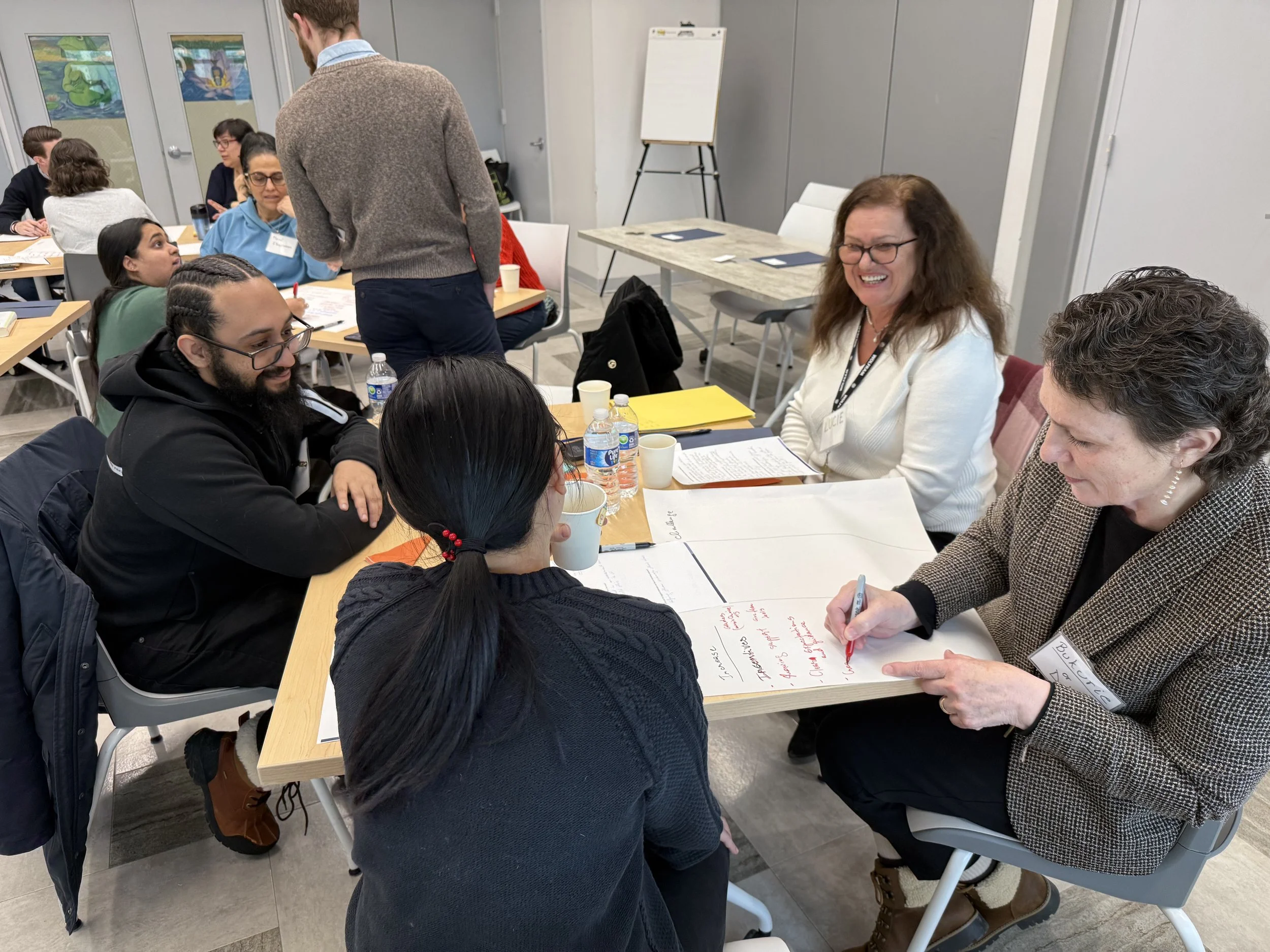 A group of diverse adults sitting around a table engaged in a collaborative activity or discussion, with some people writing and others listening, in a brightly lit room.